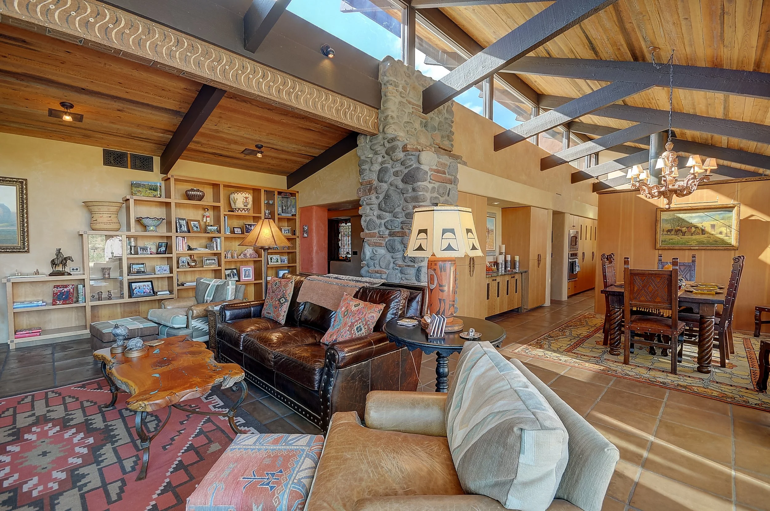 Interior of a rustic living room with wooden ceiling beams, a stone chimney, and a mix of vintage and contemporary furniture including sofas, armchairs, and a wooden dining table with chairs.