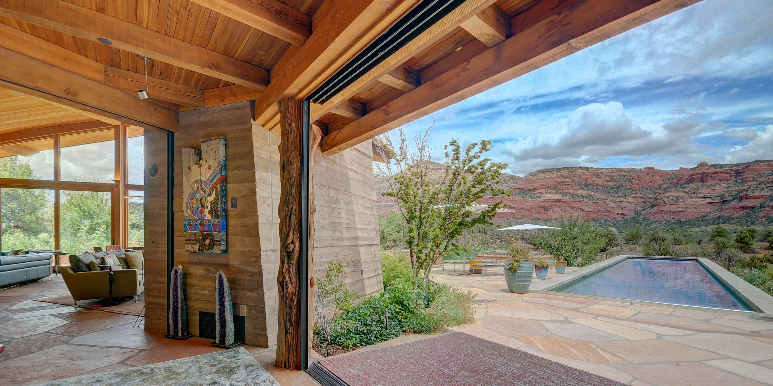 Interior view of a house with large glass doors open to an outdoor patio with a swimming pool, mountains in the background, and scattered trees and potted plants.