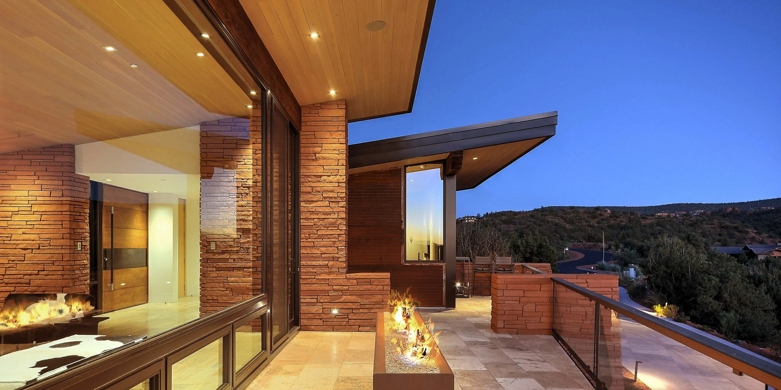 Modern house balcony at dusk with glass railing, brick walls, wooden ceiling, and a small fire feature, overlooking a mountainous landscape.
