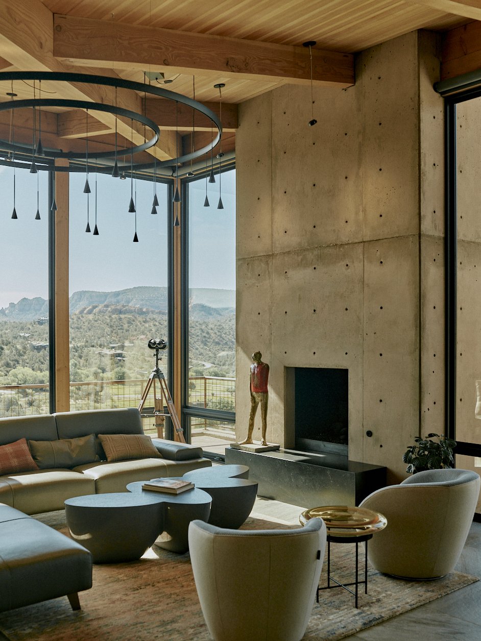 Modern living room with large floor-to-ceiling windows showing mountain views, a beige sectional sofa with throw pillows, contemporary coffee tables, and armchairs. A sculpture of a person stands near the fireplace, and a telescope is set up near the window. The ceiling features wooden beams and a circular chandelier with hanging lights.
