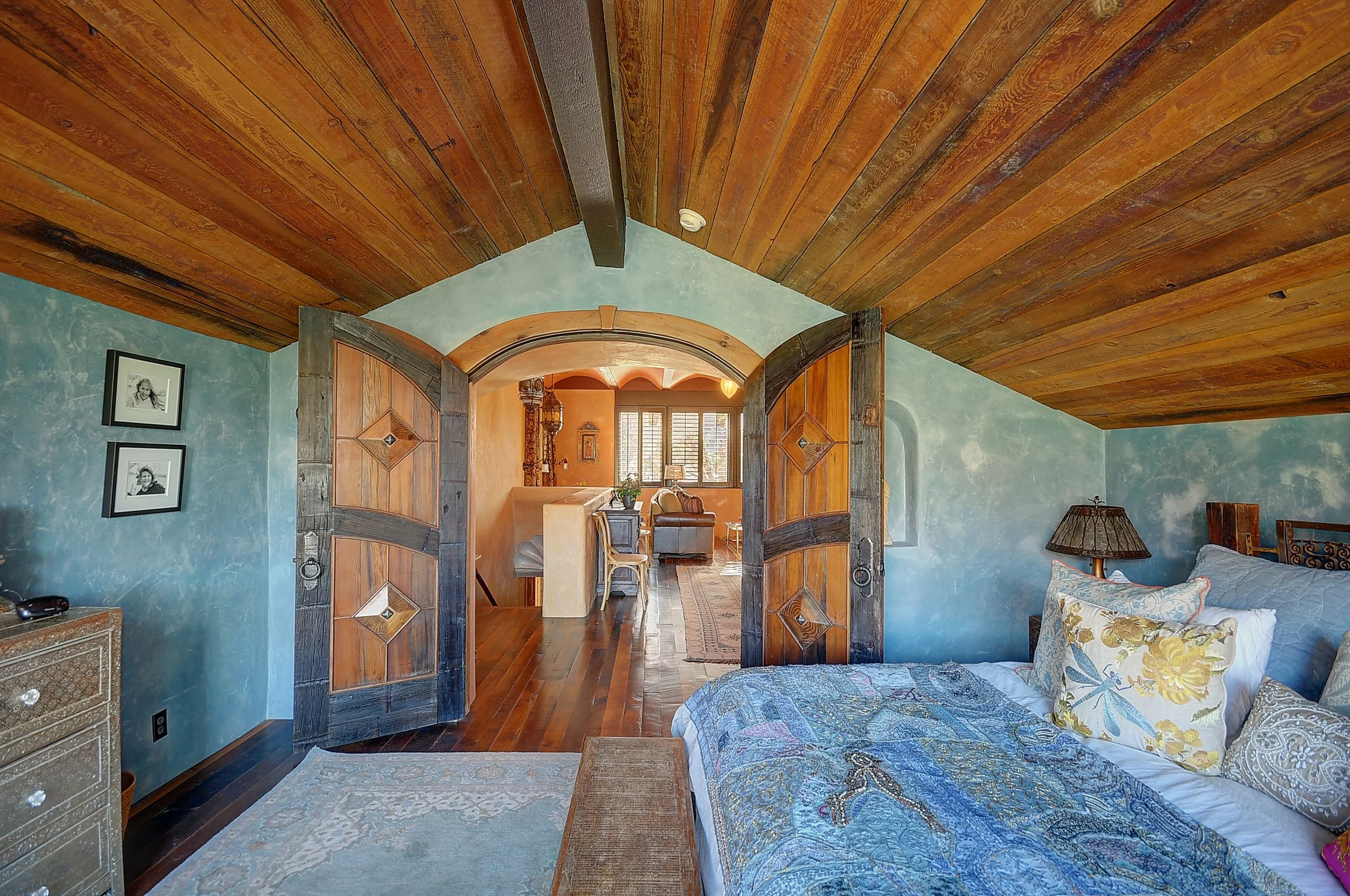 A cozy bedroom with a sloped wooden ceiling, blue textured walls, a bed with floral and patterned pillows, a lamp, and a window. French doors with dark wood framing open to a living area with wooden furniture, a window with shutters, and warm lighting.