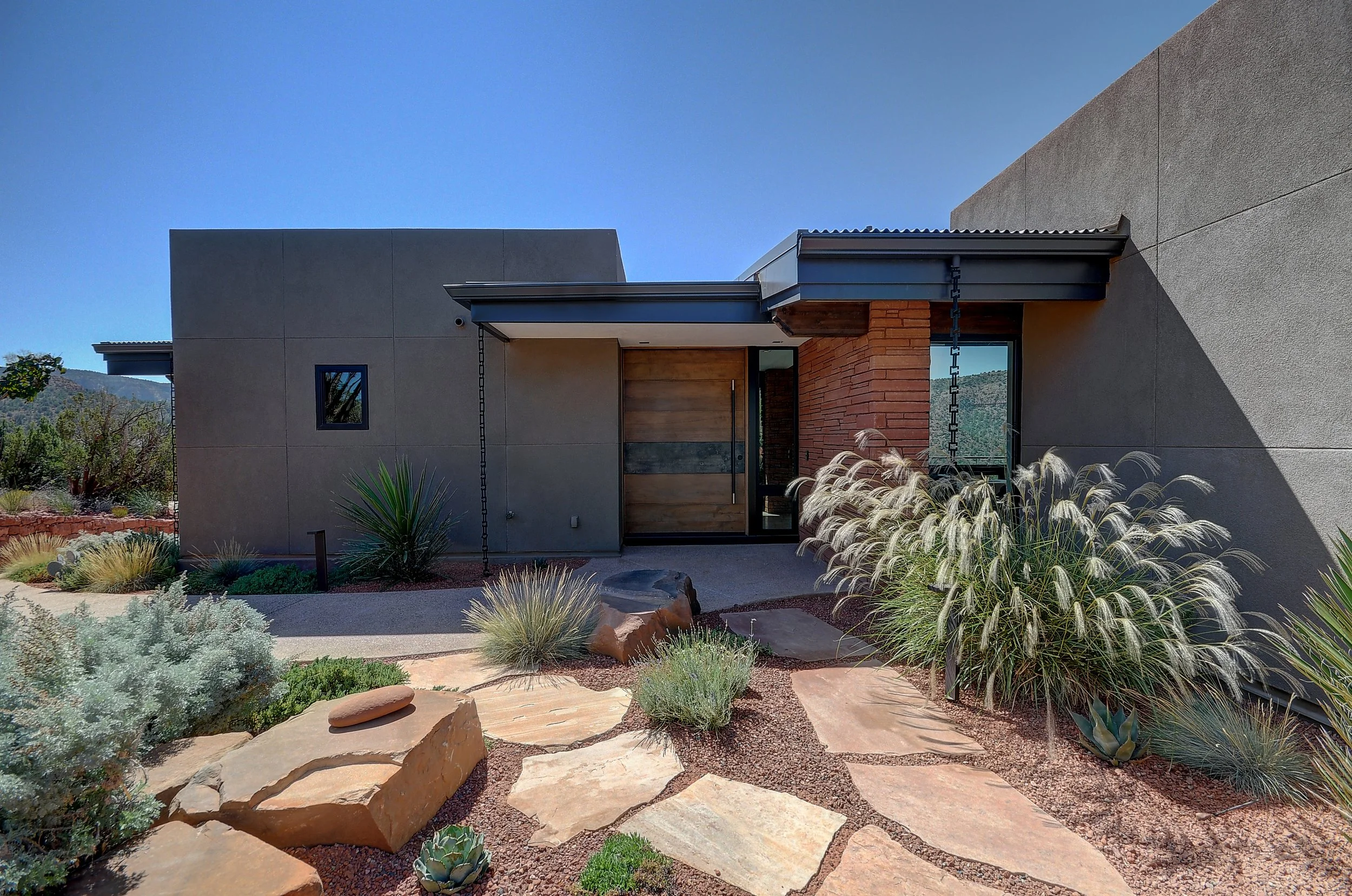 Contemporary home exterior with large pivot door and grey adobe walls, with a flagstone pathway approaching the door.