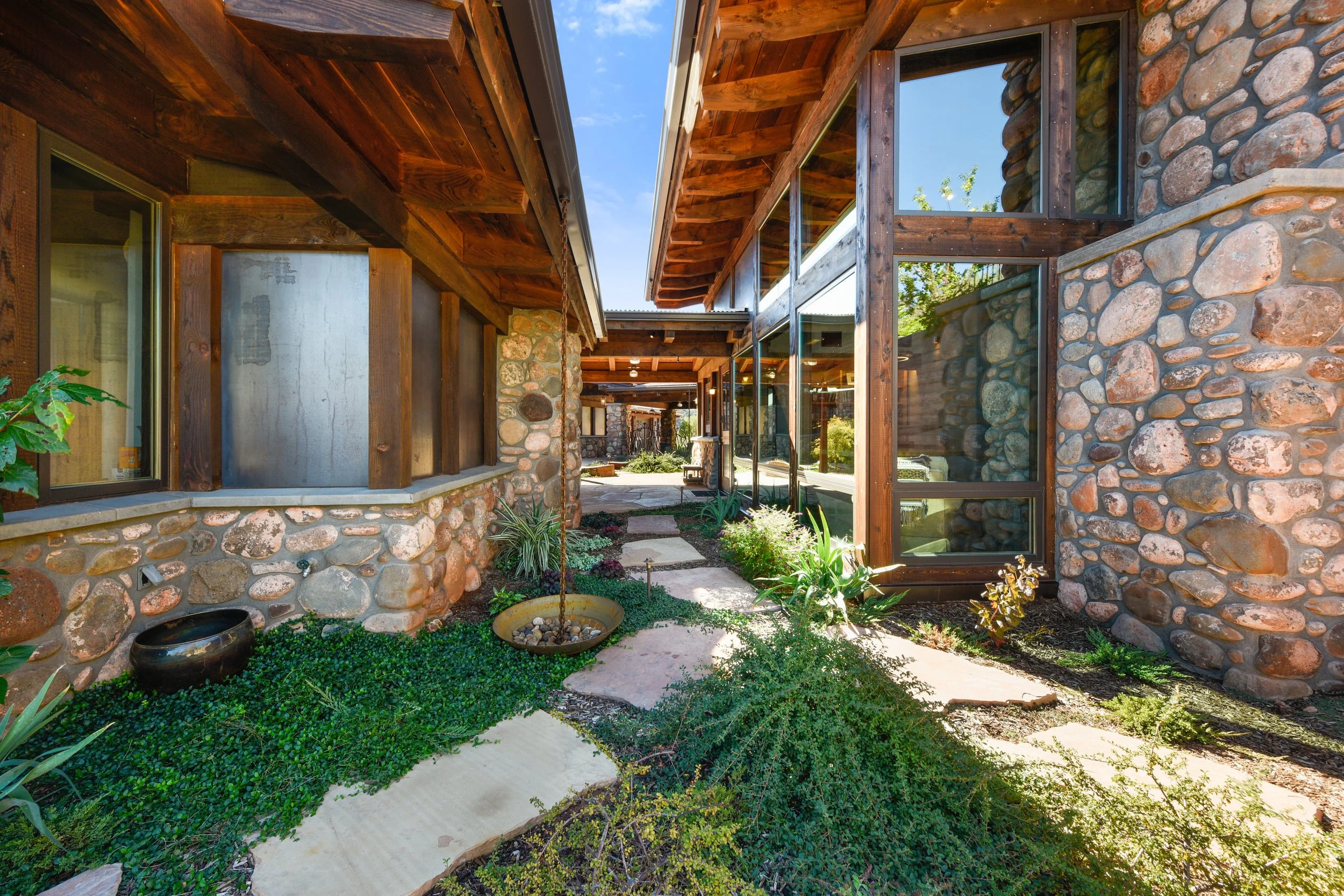 Side view of a house with a stone exterior, large windows, and wooden roof structures. There is a garden with stepping stones and various green plants in front.