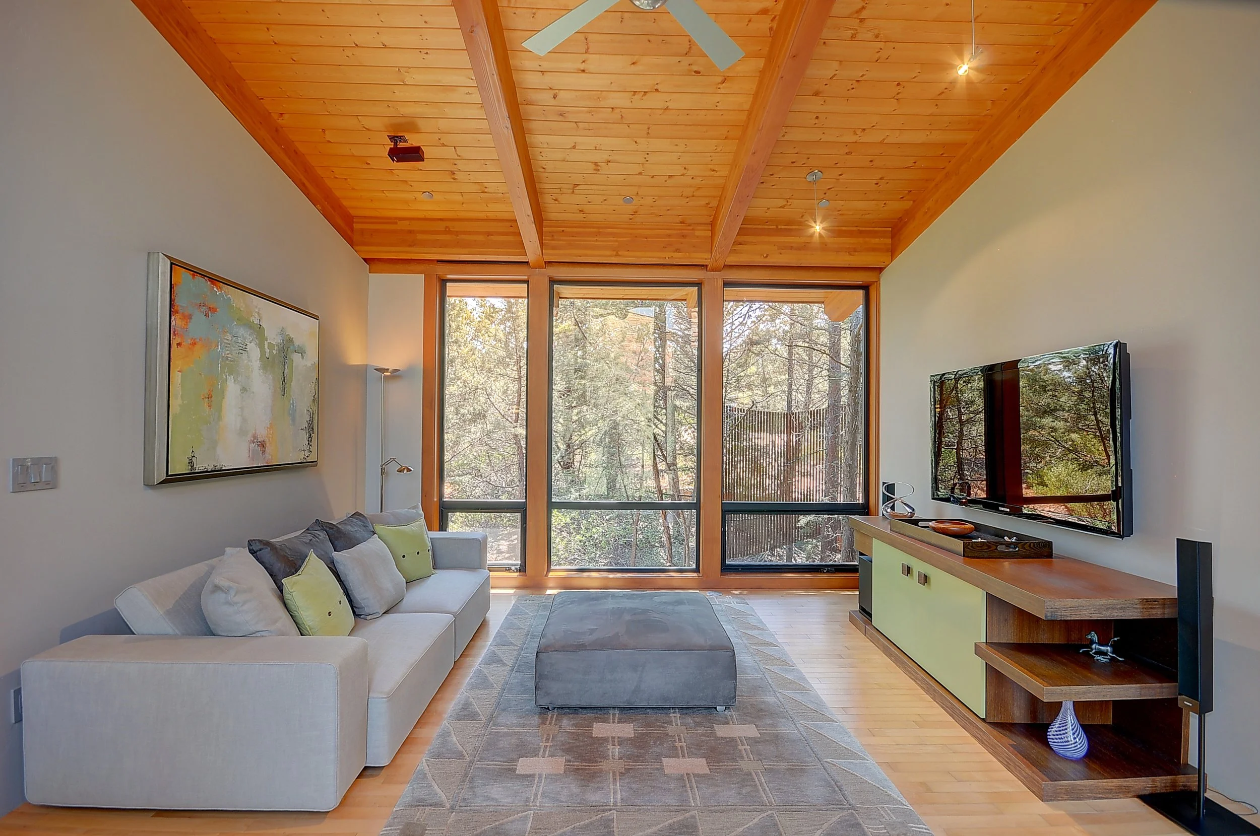 Living room with a beige sofa with pillows, a gray ottoman, a patterned rug, a wooden entertainment center, a wall-mounted TV, a large window showing trees outside, wood vaulted ceiling, and modern decor.