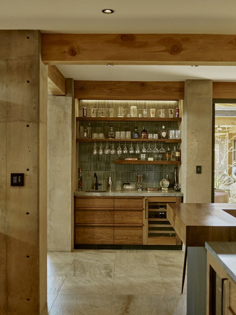 A home bar area with wooden cabinets, open shelves stocked with various liquor bottles and glassware, a small sink, and a wine cooler beneath the counter, surrounded by concrete and wooden walls with track lighting.