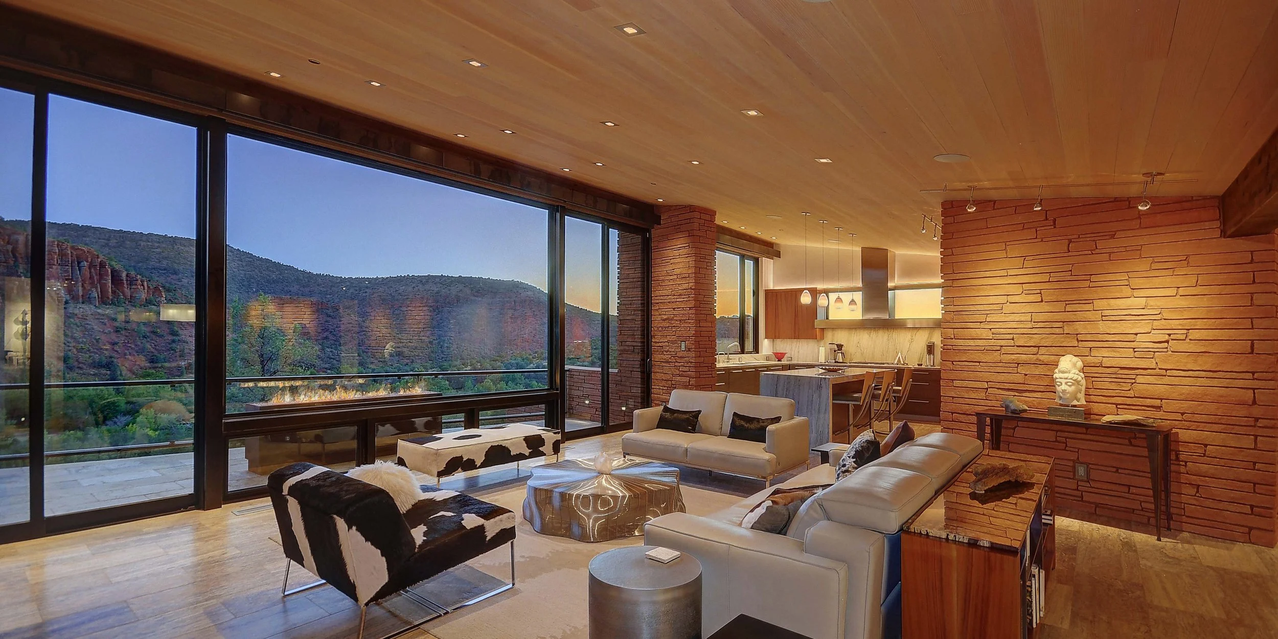 Modern living room in a home with large floor-to-ceiling windows overlooking mountains, featuring a beige sofa, black-and-white cowhide chair, round coffee table, and a brick accent wall with a sculpted head statue.