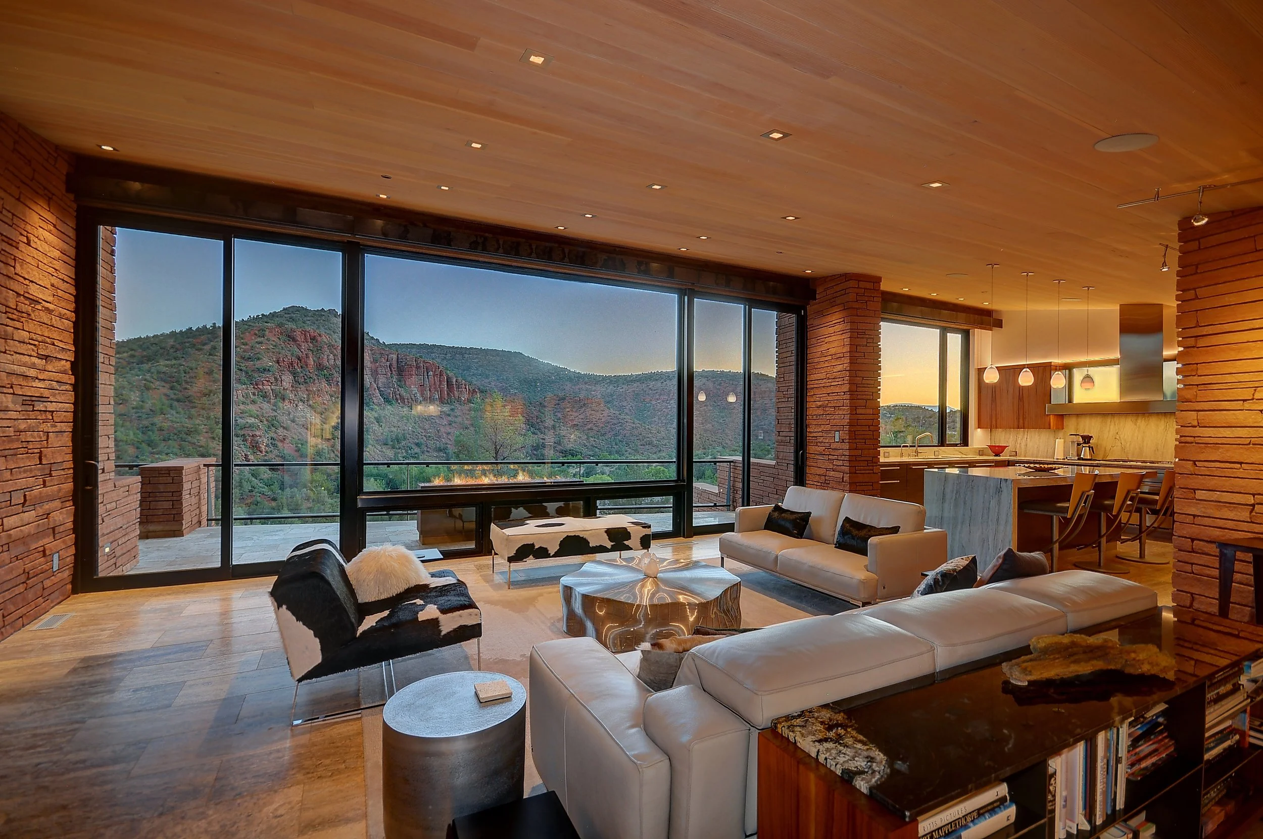 Modern living room with large floor-to-ceiling windows showing mountain views, furnished with white sofas, a black and white cowhide chair, and a gold-toned coffee table, with a wooden ceiling and kitchen area in the background.