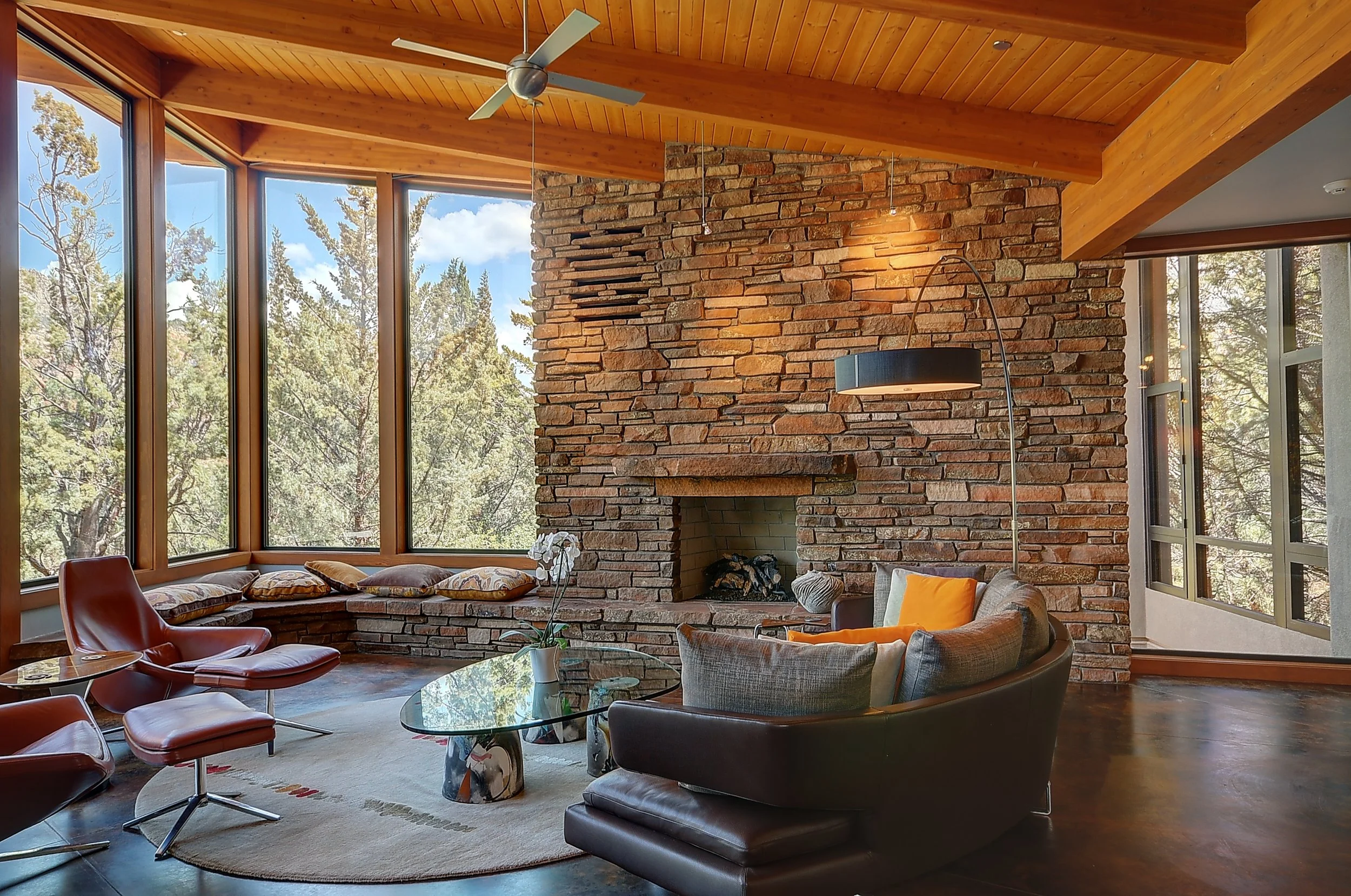 Contemporary living room with large windows, a large flagstone fireplace, modern chairs, a glass coffee table, and beautiful tongue and groove wood ceiling.