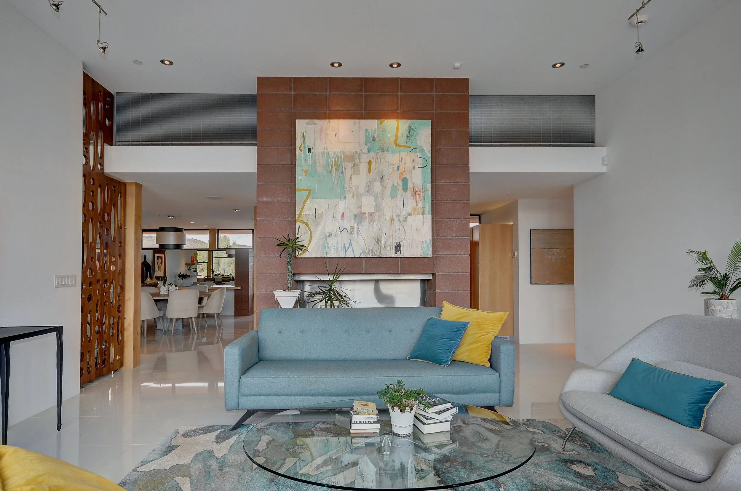 Modern living room with light blue and gray sofas, colorful pillows, a glass coffee table with books and plants, a large abstract painting above a fireplace with a brick surround, contains potted plants, a rug, and visible dining area in the background with natural light.