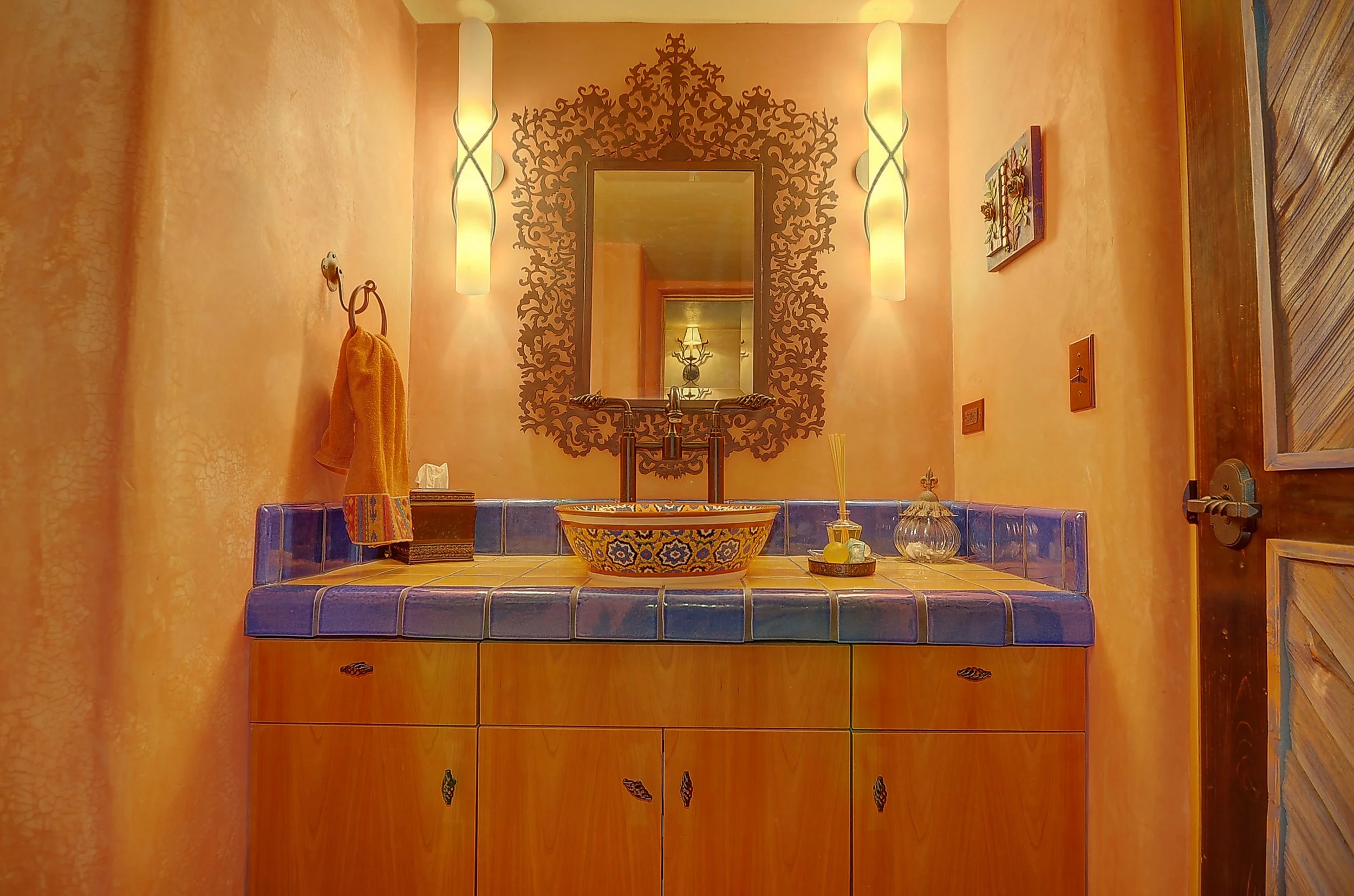 A bathroom vanity with a colorful tile vessel sink, blue tiled countertop, and wooden cabinet below. A decorative mirror with an ornate frame hangs above the sink, with two vertical wall sconces on either side. The walls are painted a warm peach color, and a hand towel hangs on a hook to the left.