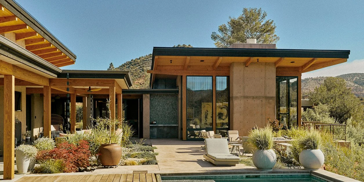 Modern house with large windows and wooden accents, outdoor patio with lounge chairs and potted plants, surrounded by trees and mountains in the background.