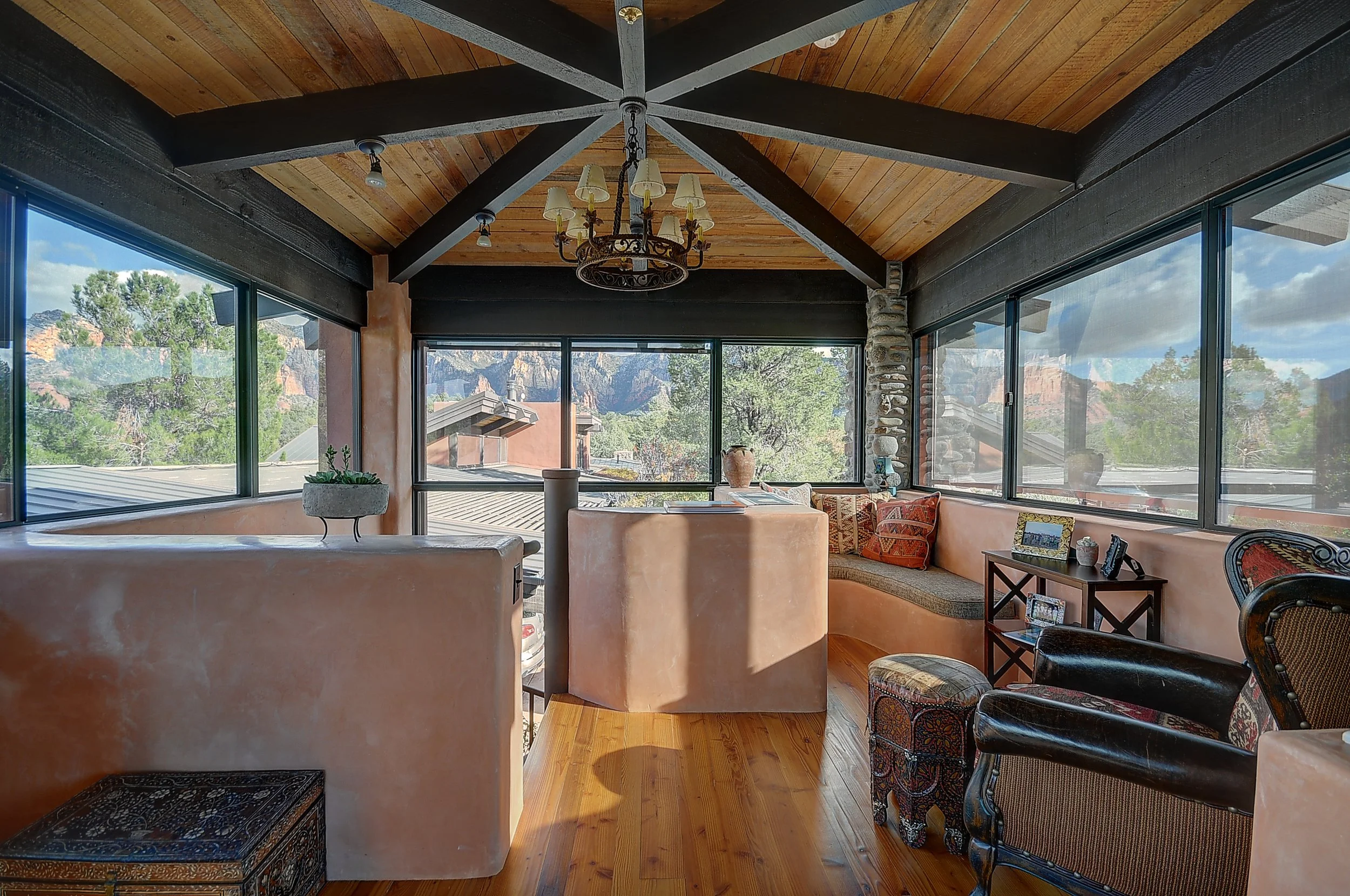 Sunroom with large windows showing mountain views, wooden ceiling with exposed beams, a chandelier, built-in bench with pillows, upholstered chairs, a small table with picture frames, and potted plants.