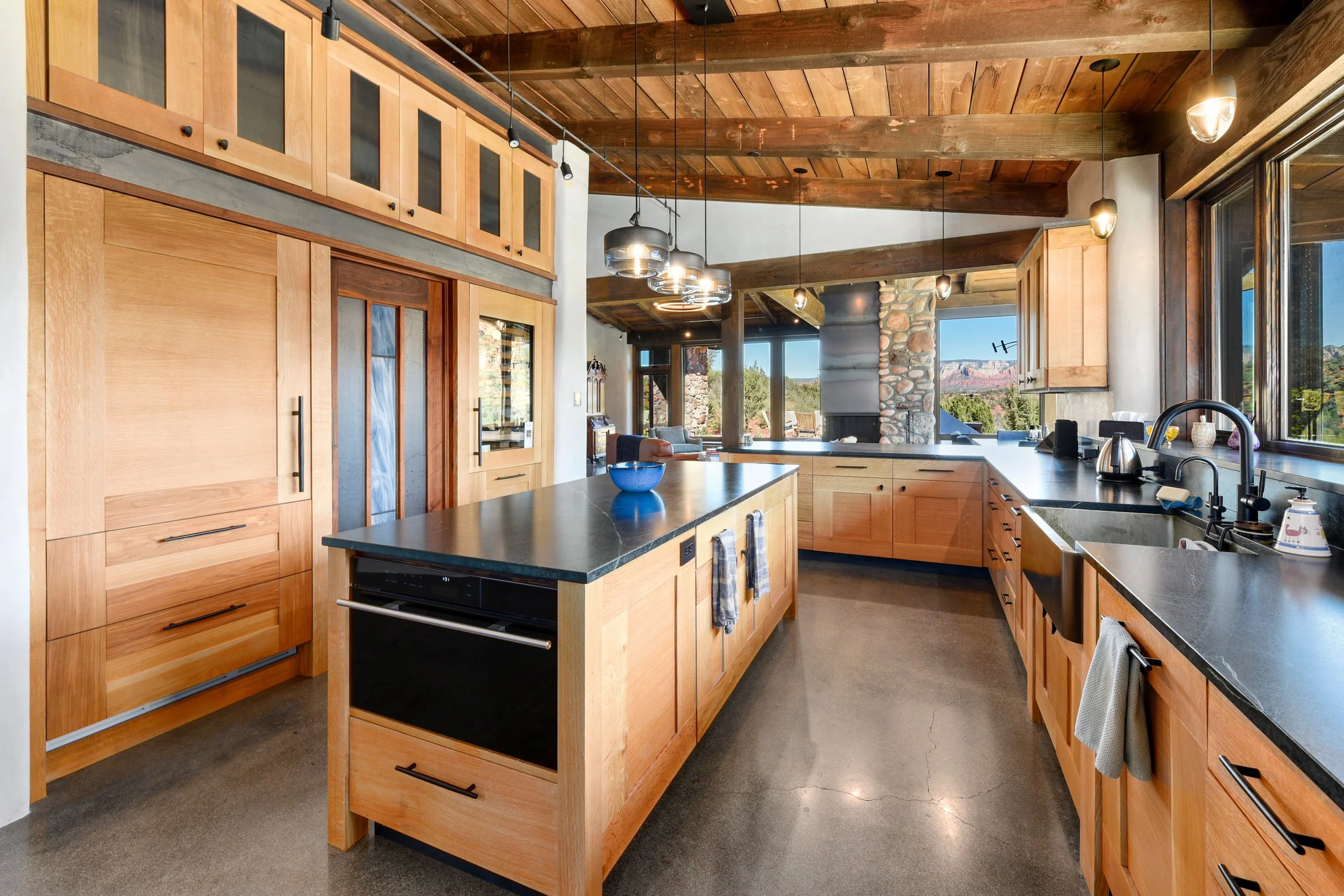 Modern kitchen with wooden cabinets, black countertops, large windows, and a view of a stone fireplace and outdoor landscape.