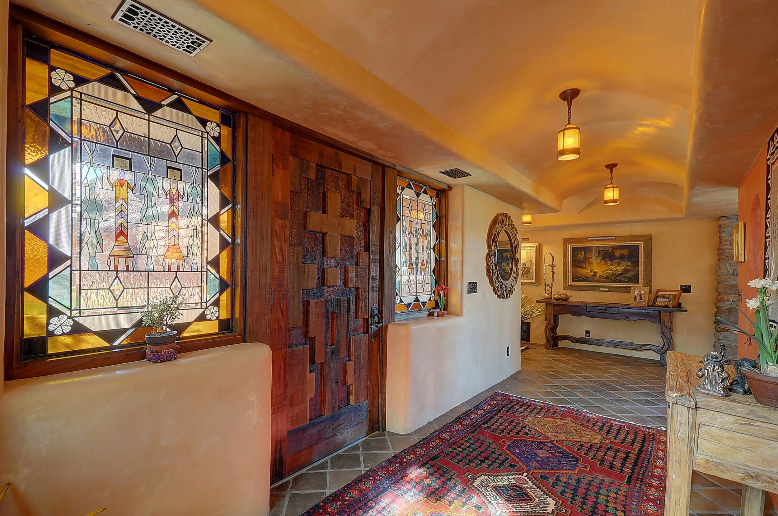Interior of a home with stained glass windows, a wooden door with geometric design, a decorative rug on a tiled floor, and rustic furniture and artwork.