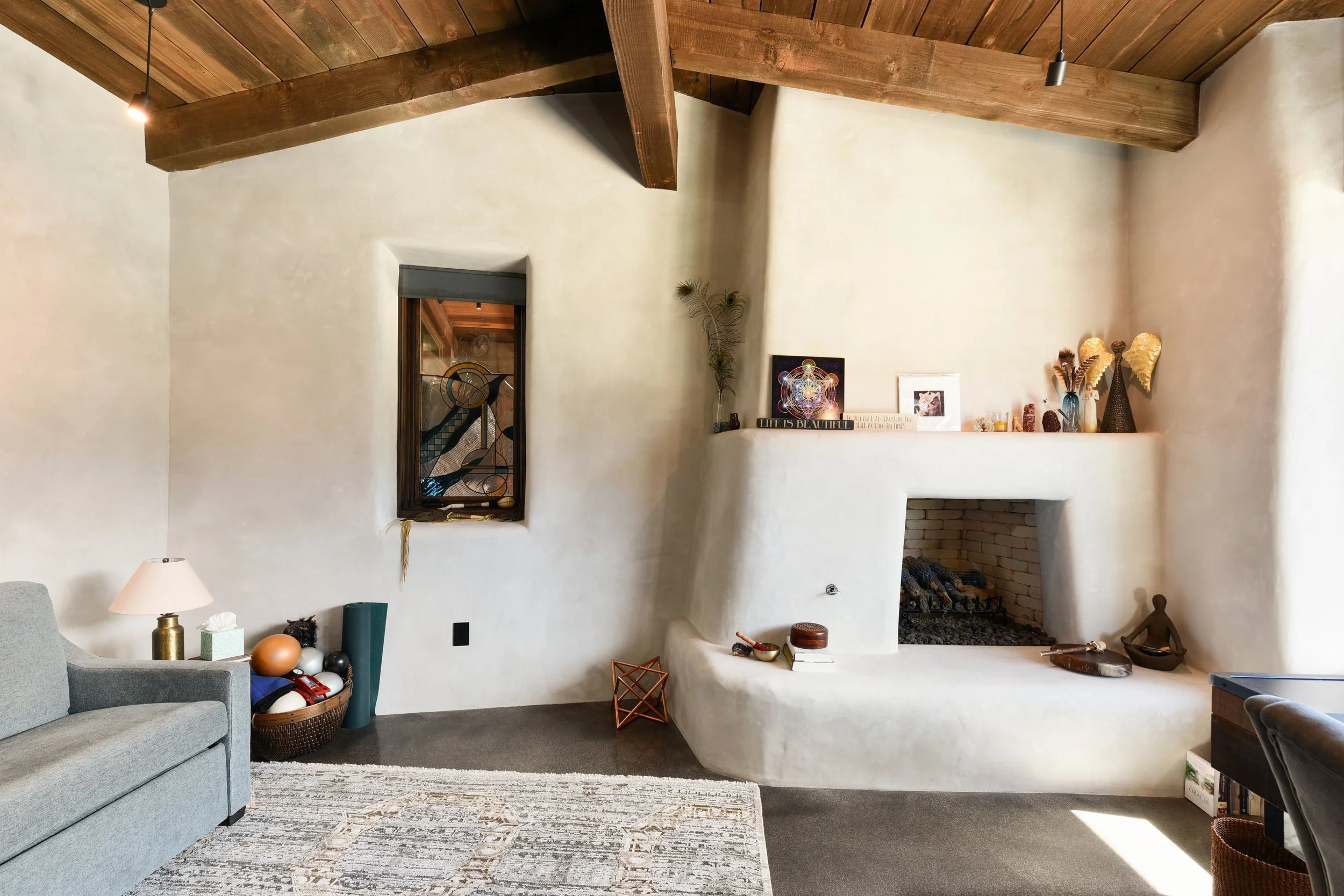 Living room with a white curved plaster fireplace, wooden ceiling beams, a small window with stained glass, a rustic light fixture, a gray sofa, a rug, and decorative items on the mantel and side tables.