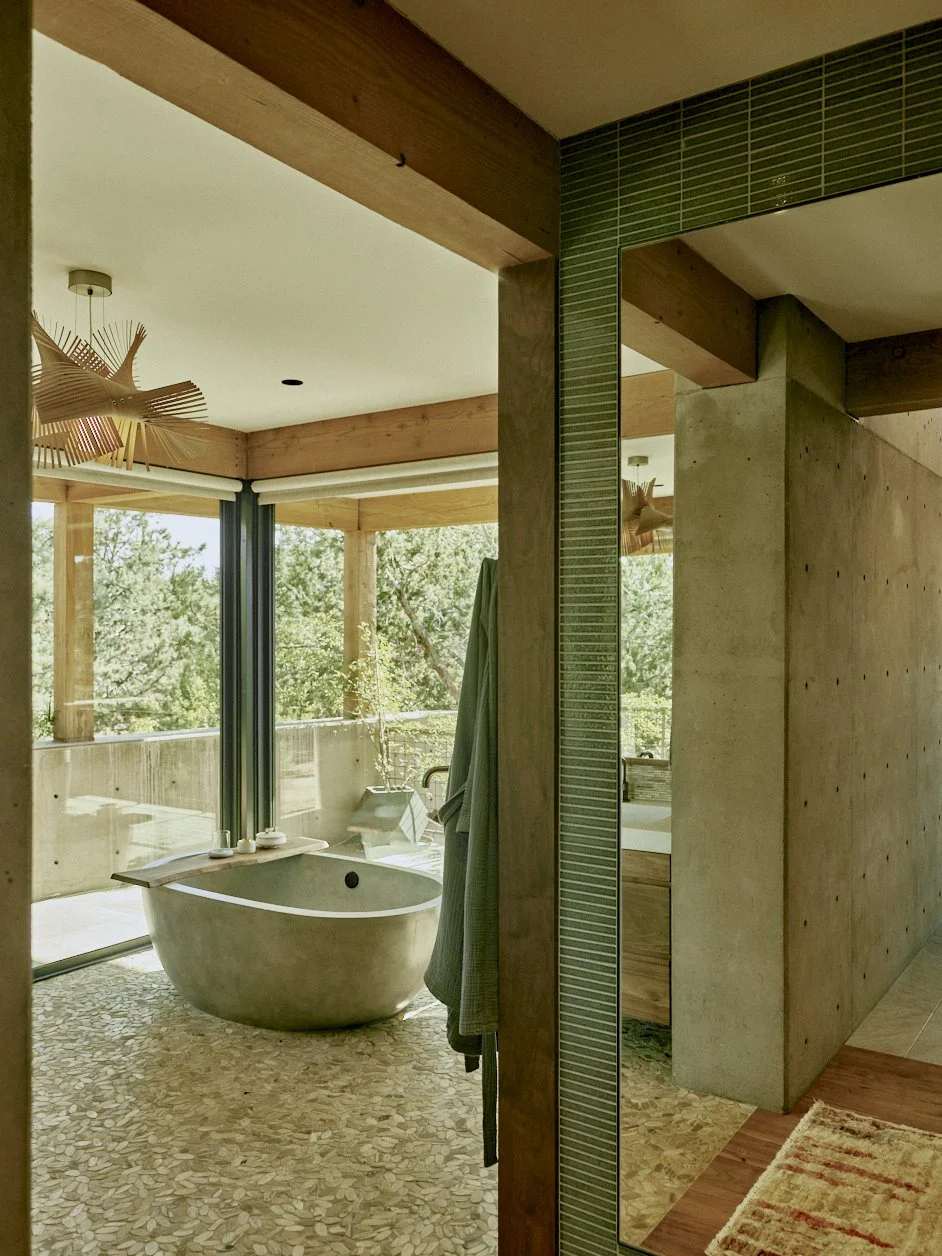 Modern bathroom with free-standing bathtub, large sliding glass doors leading to a balcony with trees outside, wood and concrete accents, and a mirror reflecting part of the room.