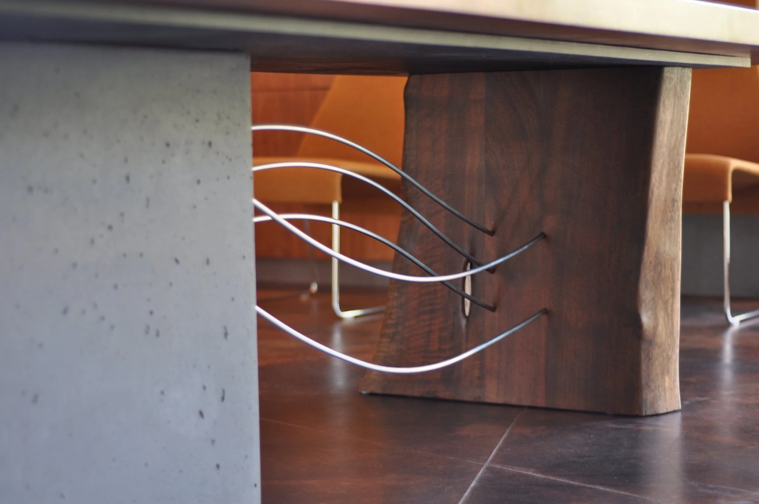 Close-up of a custom fabricated dining table featuring curved metal wires extending from a wooden leg to a concrete mass beneath the table.