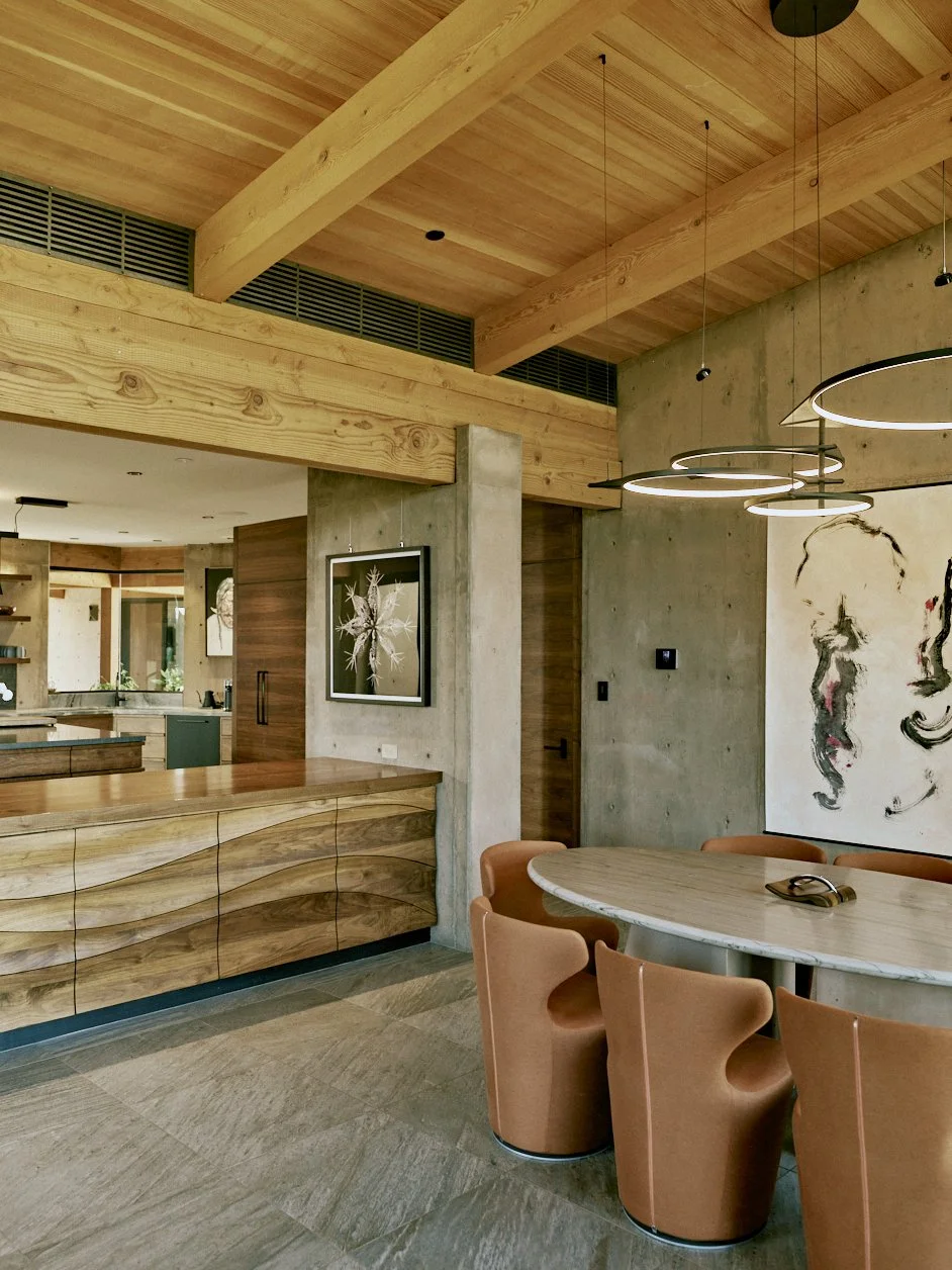 Modern dining area with a round marble table, orange upholstered chairs, wooden ceiling beams, and contemporary wall art.