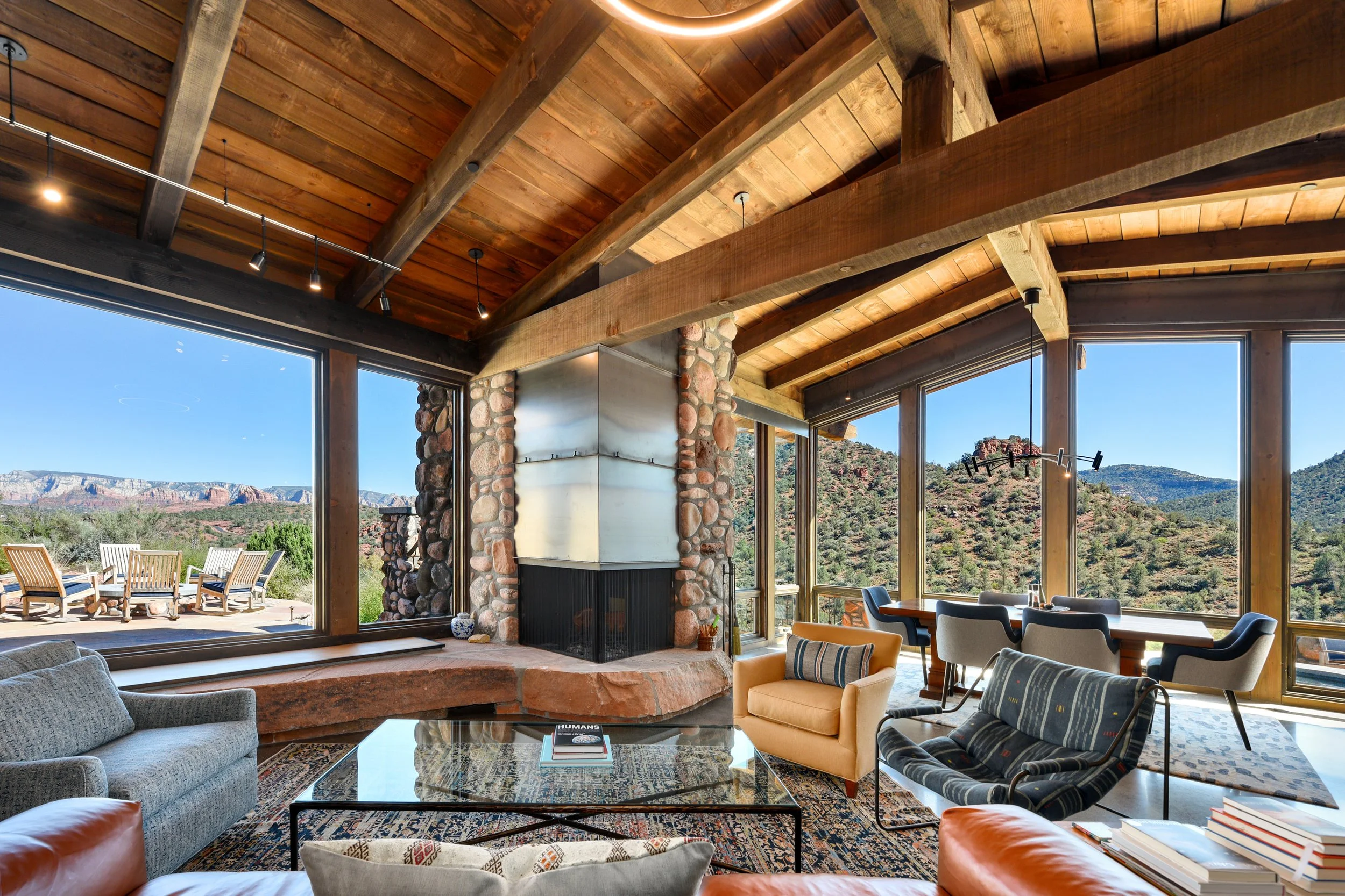 Interior of a modern mountain home living room with large windows, wooden ceiling beams, a stone fireplace, and scenic mountain views outside.