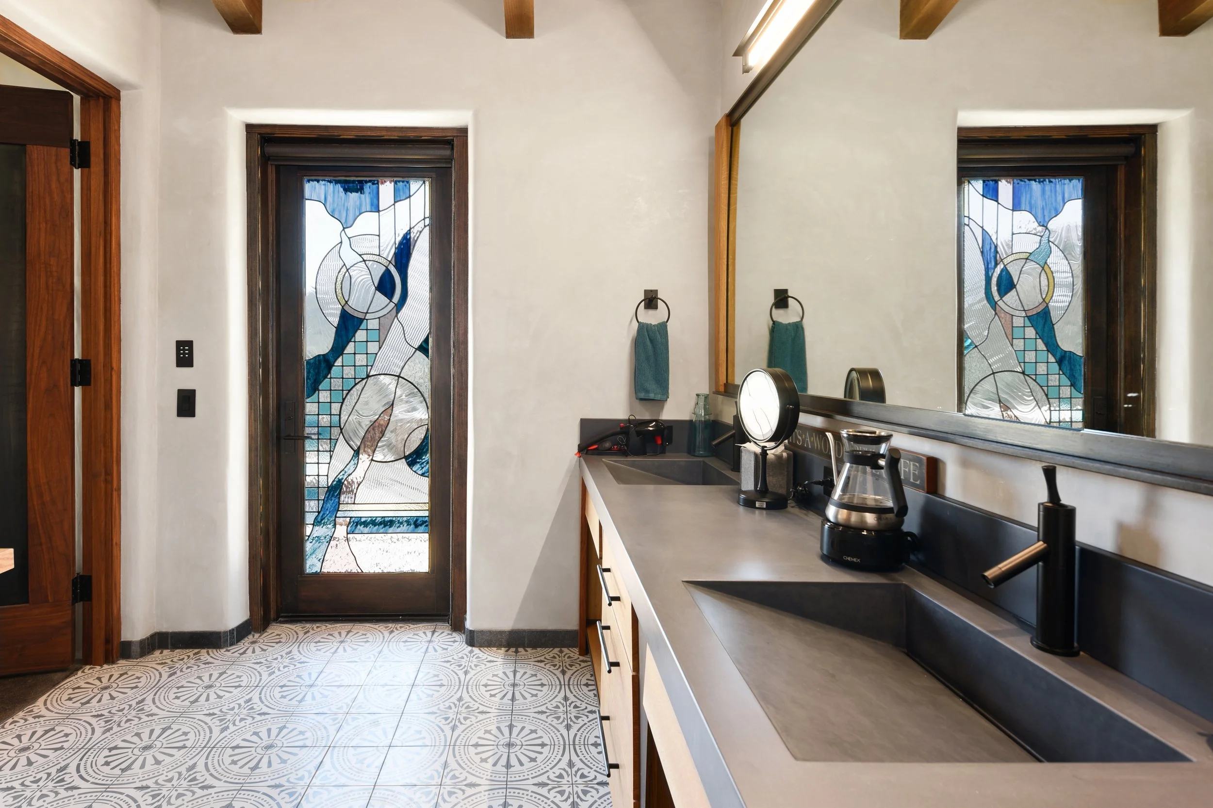 Modern bathroom with a concrete sink basin and mirror, featuring a stained custom stained glass door with abstract blue and white designs, light-colored walls, tiled floor, and wooden accents.