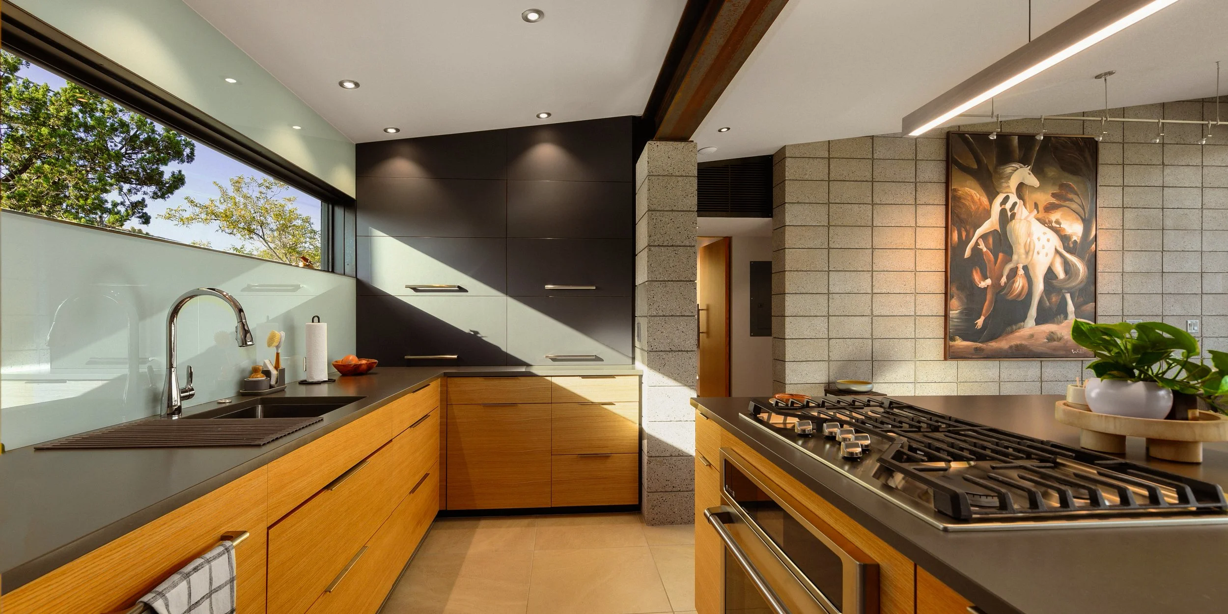 Modern kitchen with wooden cabinets, black countertops, a stove, and a large window showing trees outside. A plant is on the counter, and a painting of a unicorn with a dog on its back hangs on the brick wall.