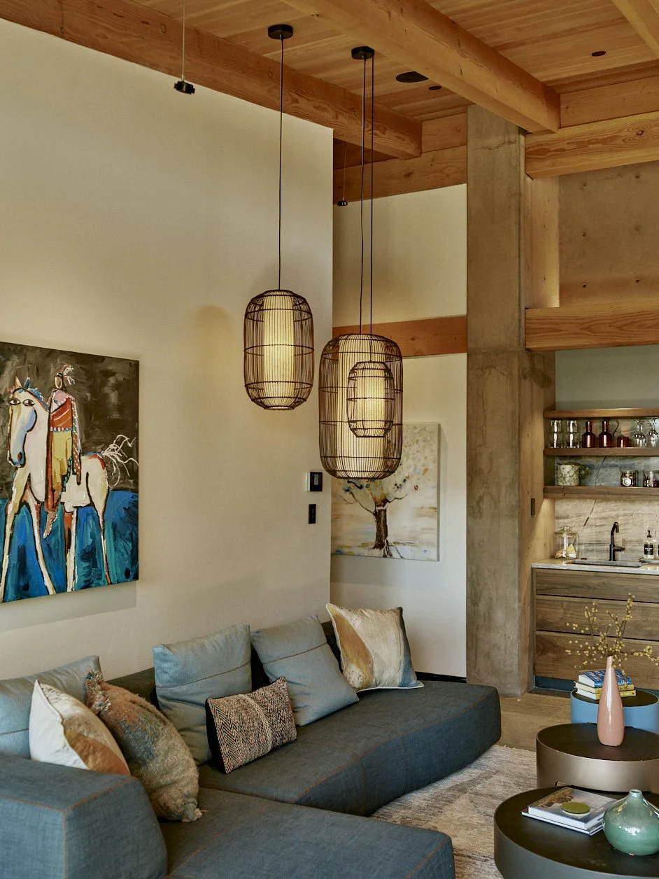 Interior of a modern living room with a dark gray sofa adorned with patterned and fur pillows, wooden ceiling beams, three hanging woven pendant lights, and contemporary wall art. There's a small round coffee table with a pink vase and books, and a corner kitchen with open shelves and a concrete column.