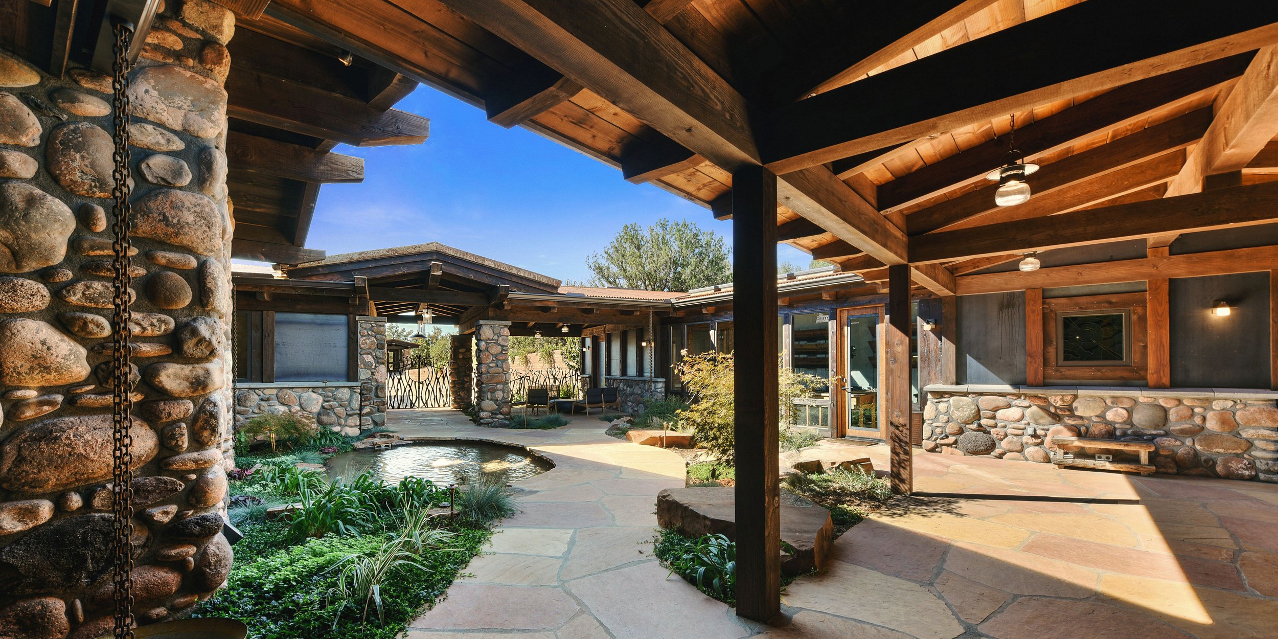 Outdoor patio area with stone walls, a pond, and wooden beams overhead, under a clear blue sky.