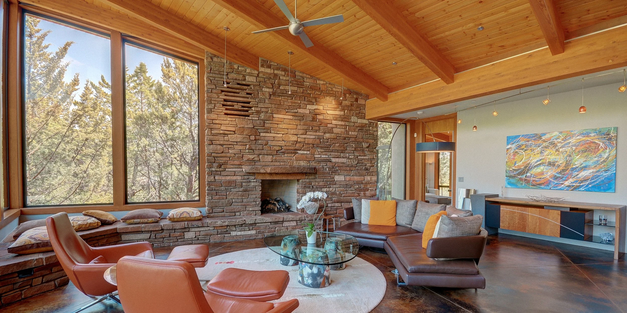 Living room with large windows, brick fireplace, wooden ceiling, modern furniture, and abstract artwork.
