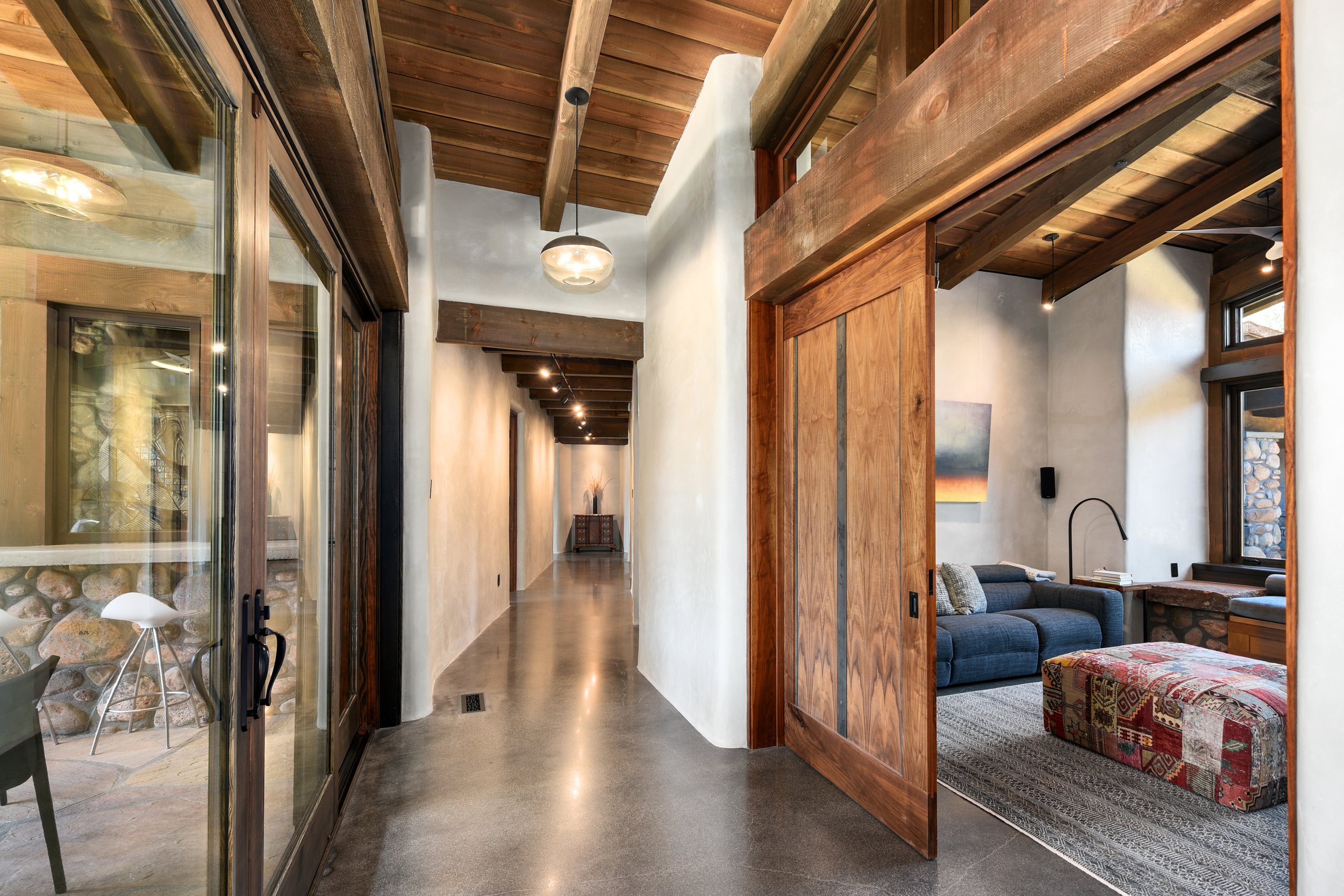 Interior of a modern home with a hallway, a room with a blue sofa, a wooden sliding door, and large windows, featuring wood and concrete finishes.