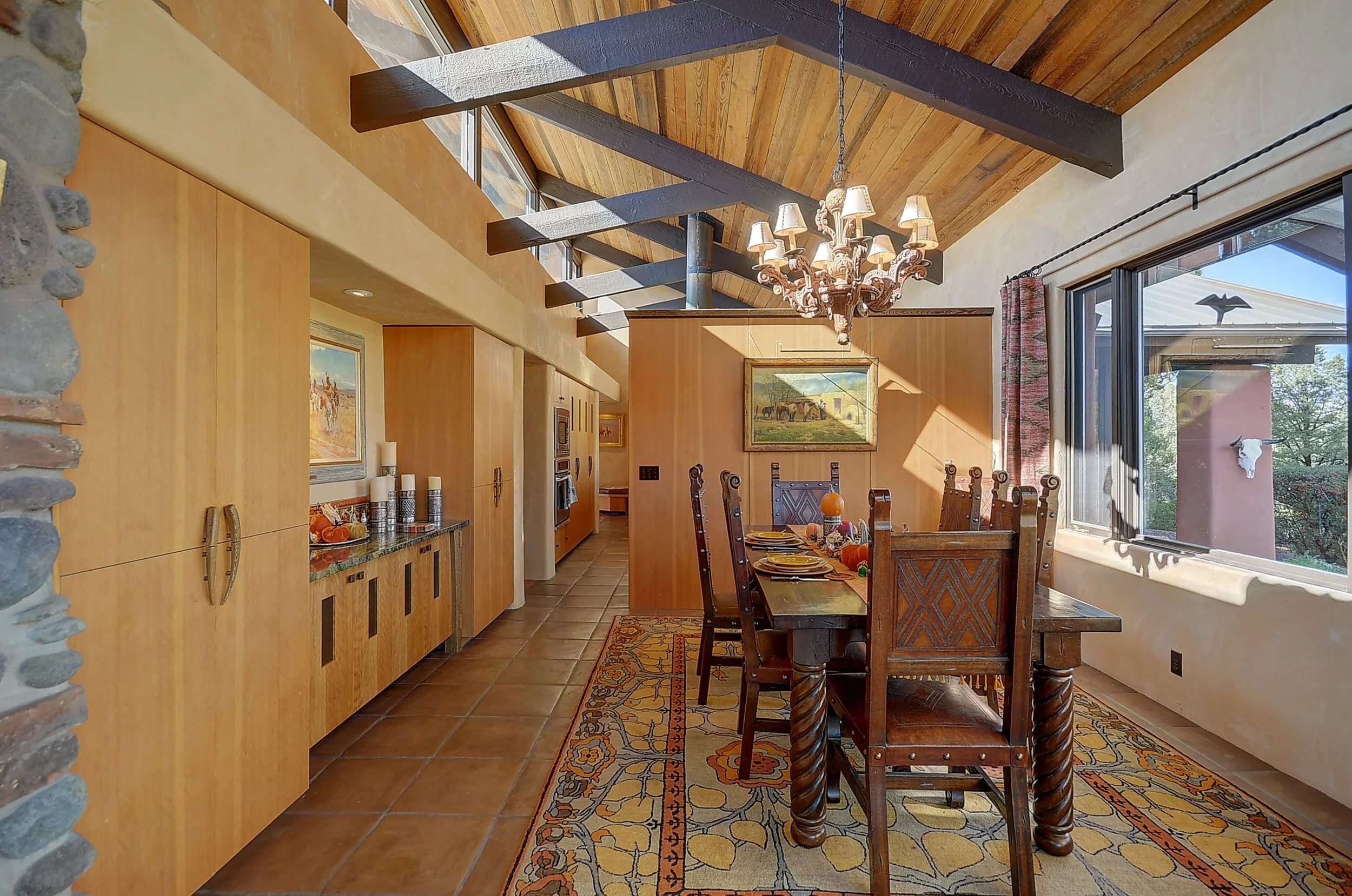 A cozy dining room with a dark wood dining table and matching high-back chairs, a large window with a view of trees, a patterned area rug, a chandelier hanging from a wooden ceiling with exposed beams, and a built-in wooden cabinet with decorative items and artwork on the walls.