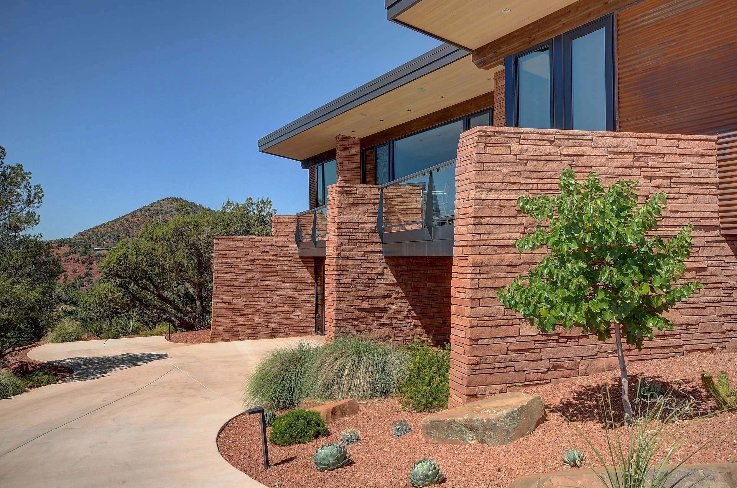 Contemporary adobe house with brick and wood exterior, large glass windows, a small balcony with metal railing, landscaped garden with small plants and a young tree, and mountain scenery in the background.