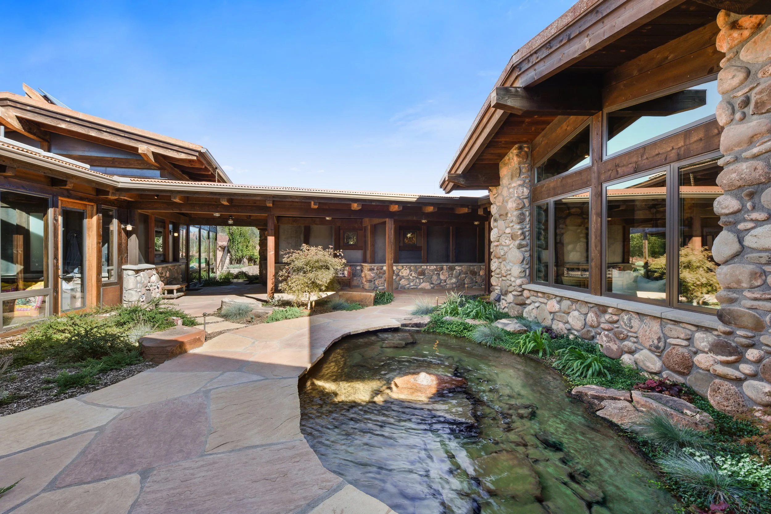 View of a craftsman house with large windows, flagstone walls and exposed timber, surrounding a small pond with lush plants, under a clear blue sky.