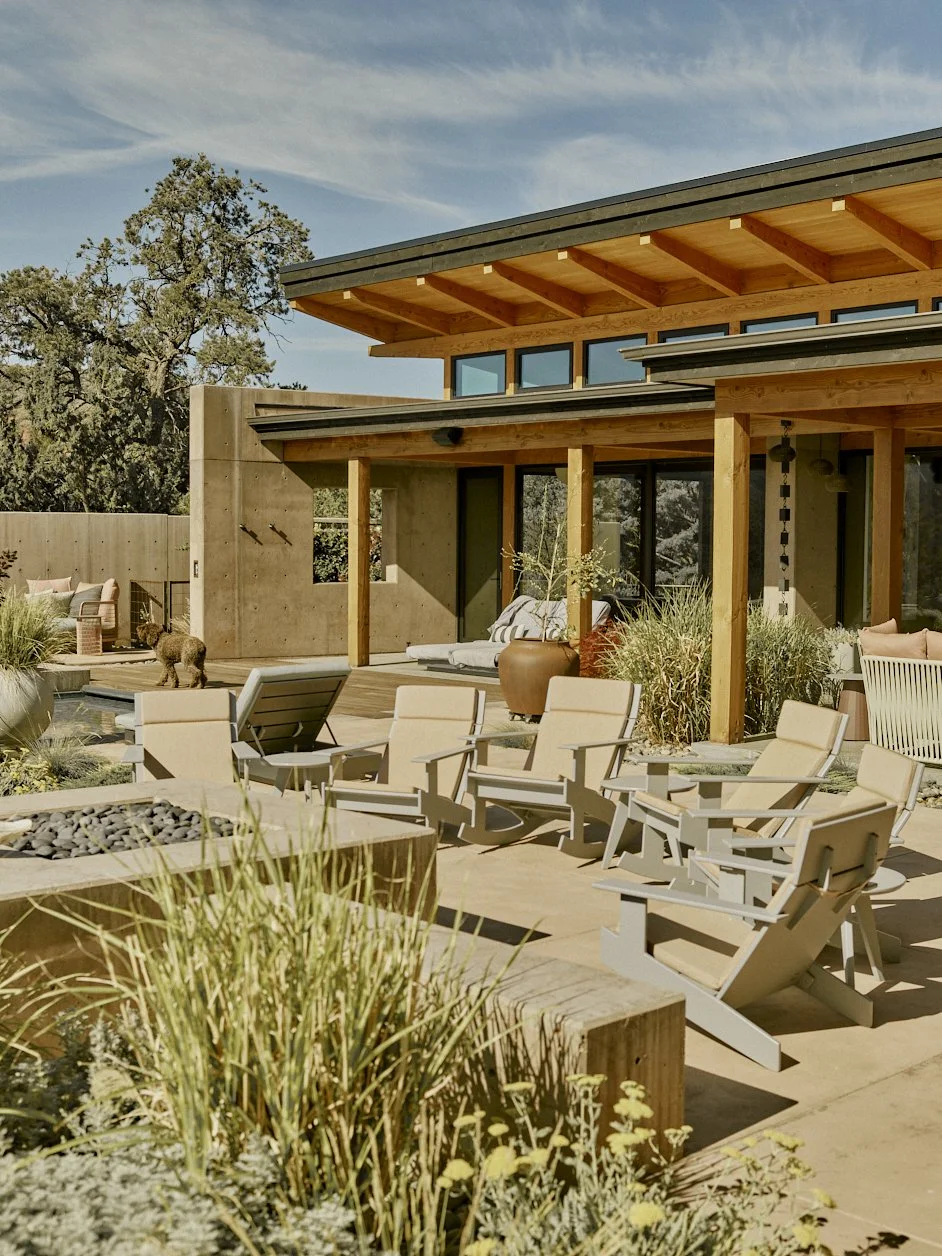 Modern house with patio area featuring beige chairs, potted plants, and a small dog, surrounded by outdoor furniture and landscape with trees and clear blue sky.