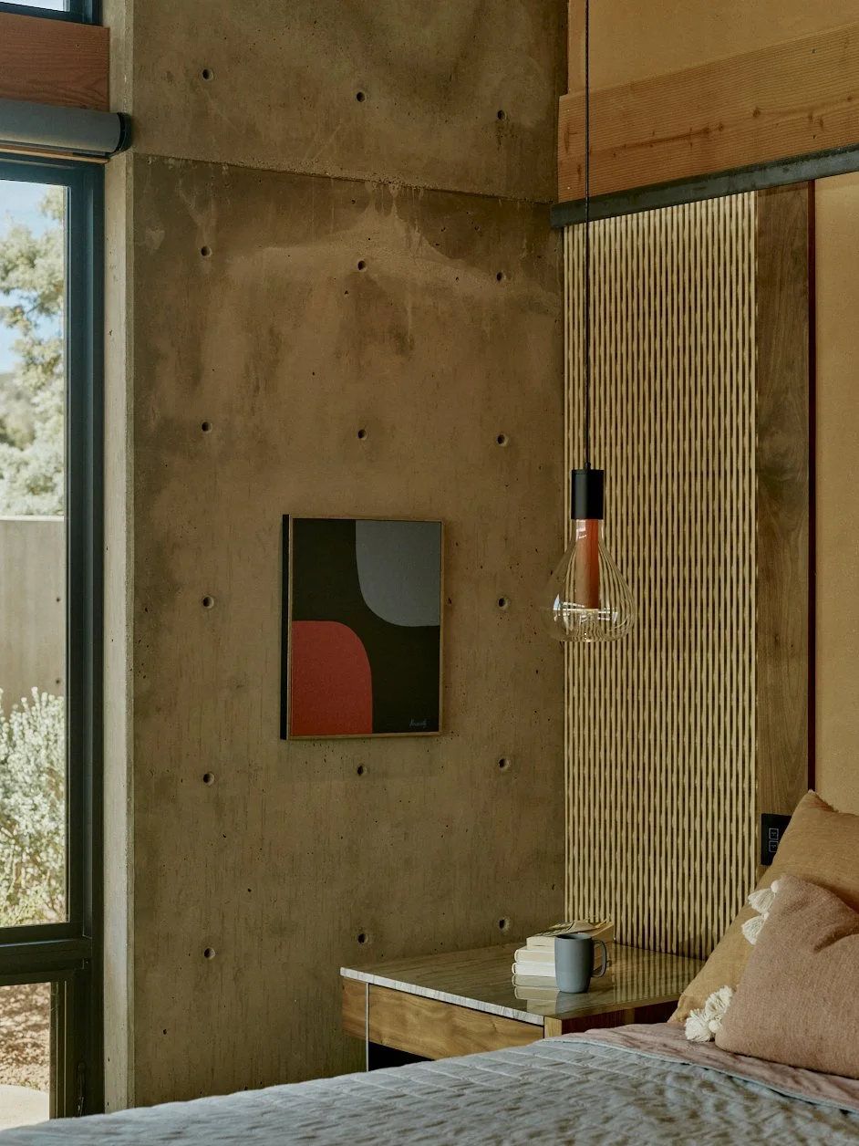 Interior of a bedroom with a concrete wall, geometric art piece, pendant light, wooden accents, a bedside table with a mug and books, and a bed with pink and beige bedding.