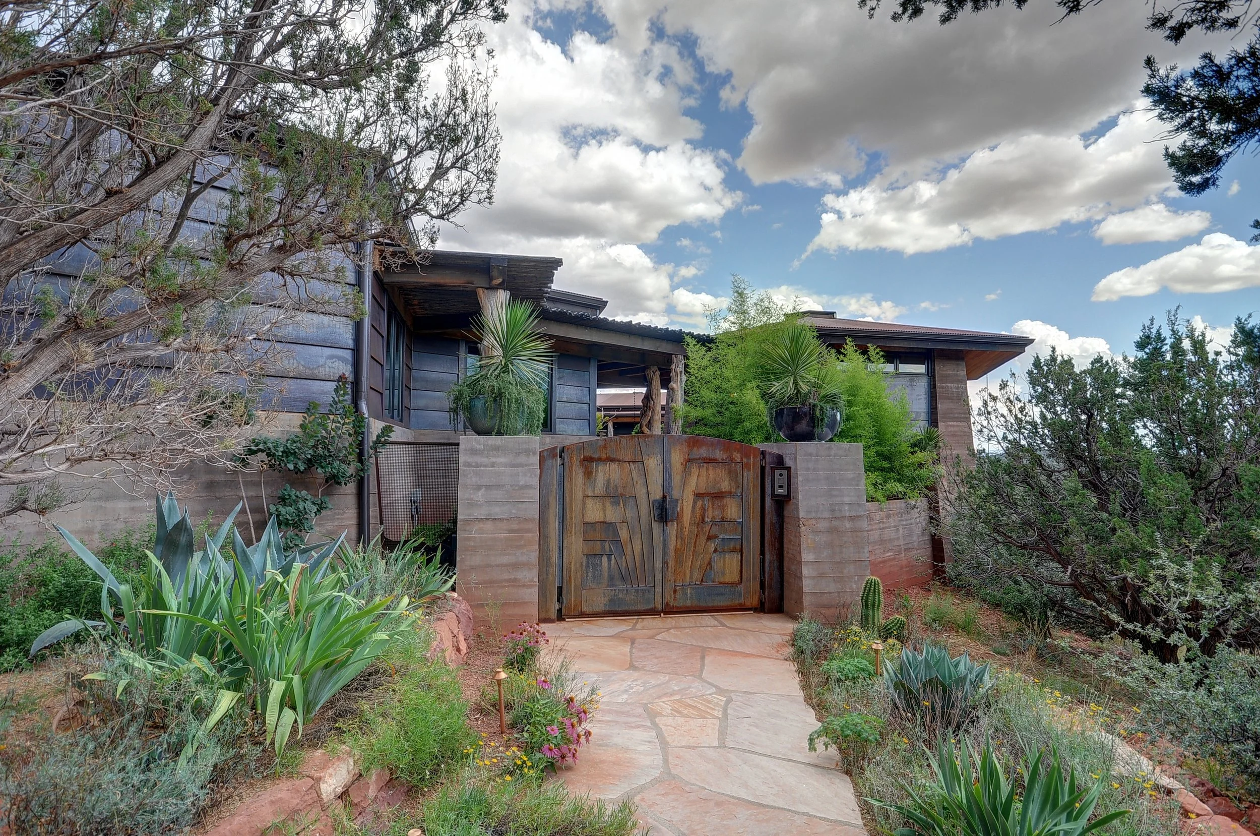 A house with a rustic metal gate, surrounded by desert-style plants including cacti and agave, set in a landscaped garden with a stone pathway, under a partly cloudy sky.