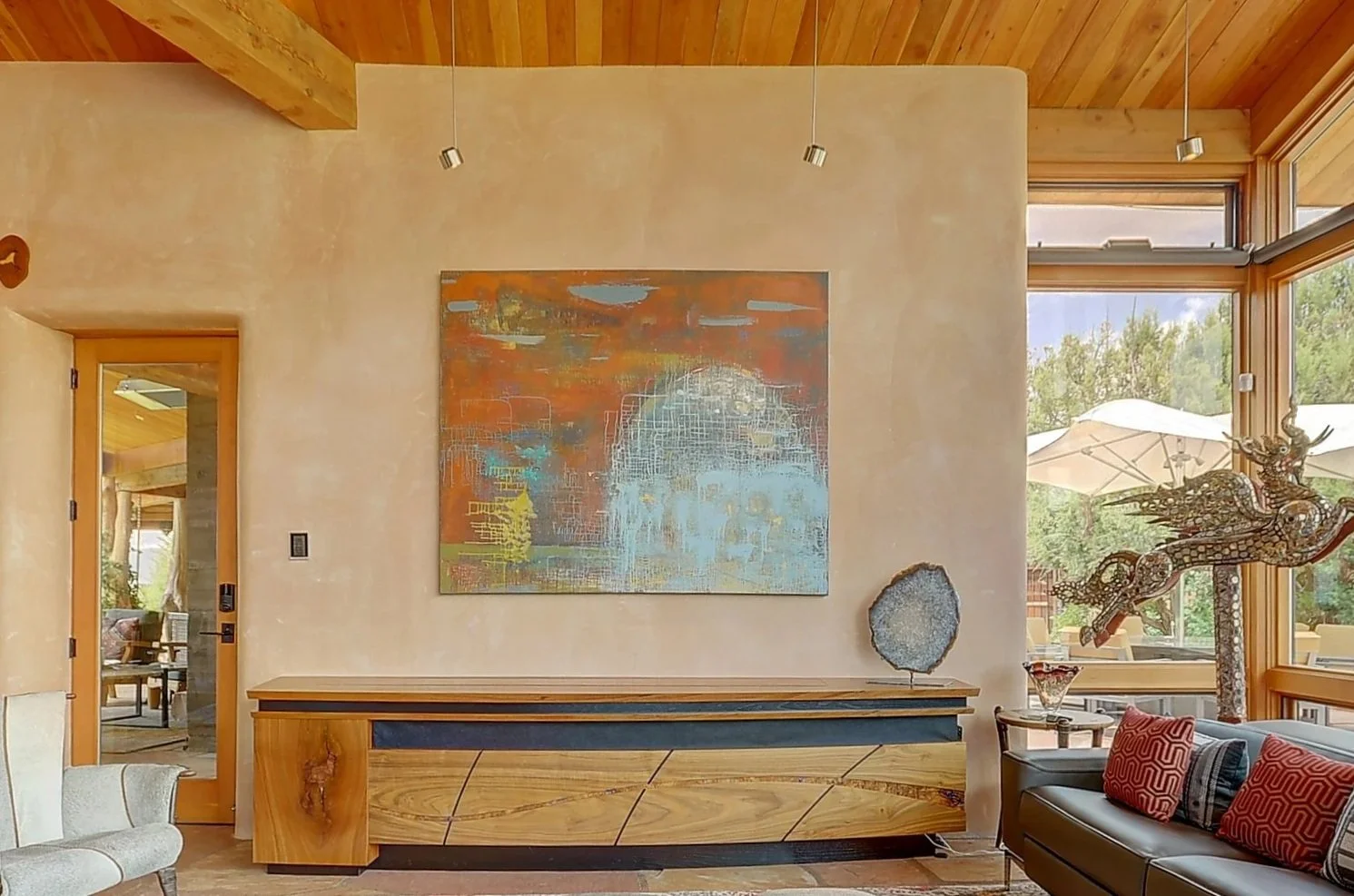 Living room with a colorful abstract painting on the beige wall, a contemporary wooden sideboard, decorative items including a geo crystal, and a dragon sculpture near large windows with patio umbrellas outside.