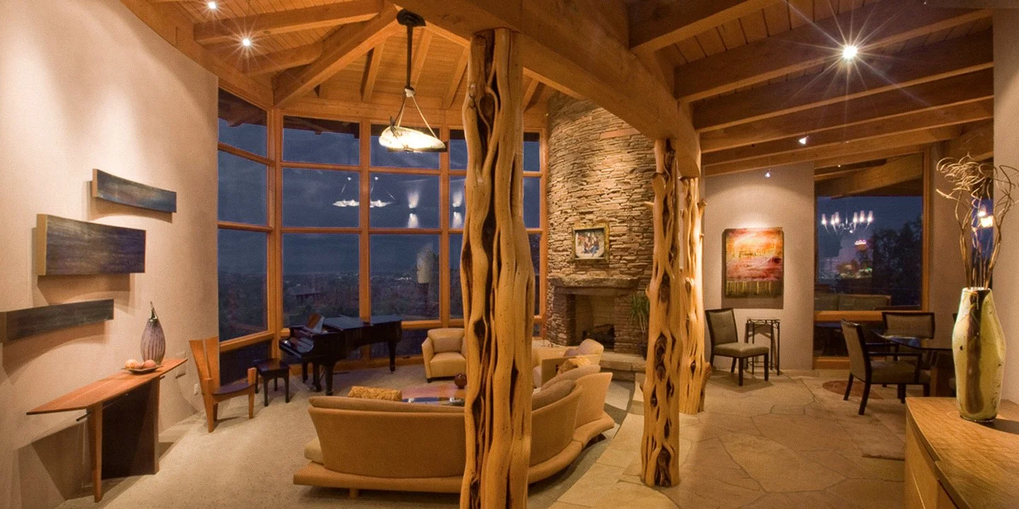 Cozy living room with wooden beams, a stone fireplace, a large window showing a night sky, a grand piano, armchairs, and decorative vases.