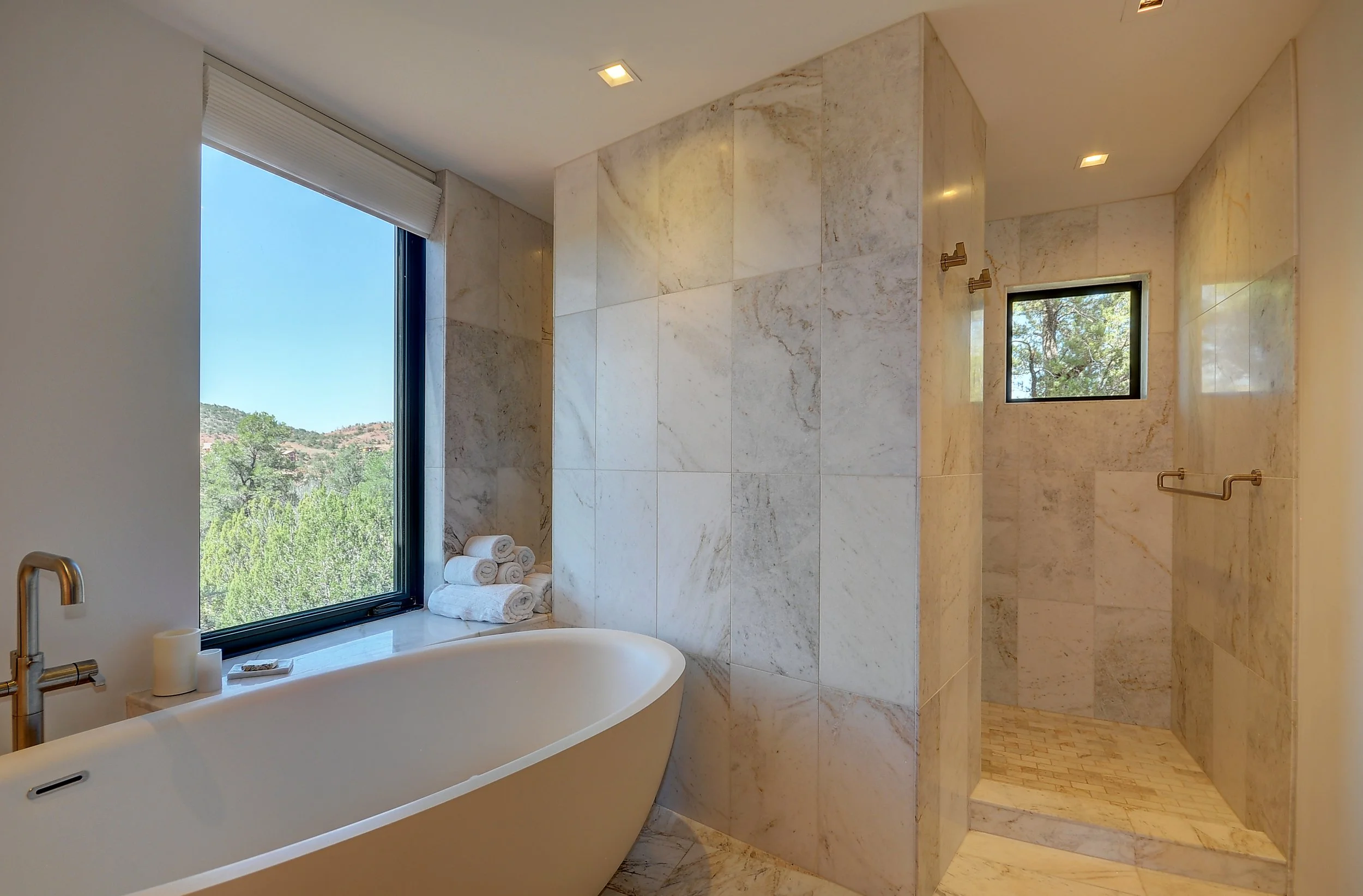 Modern bathroom with a freestanding bathtub near a large window with a view of trees and hills, and a separate shower area with marble tiles and a small window.