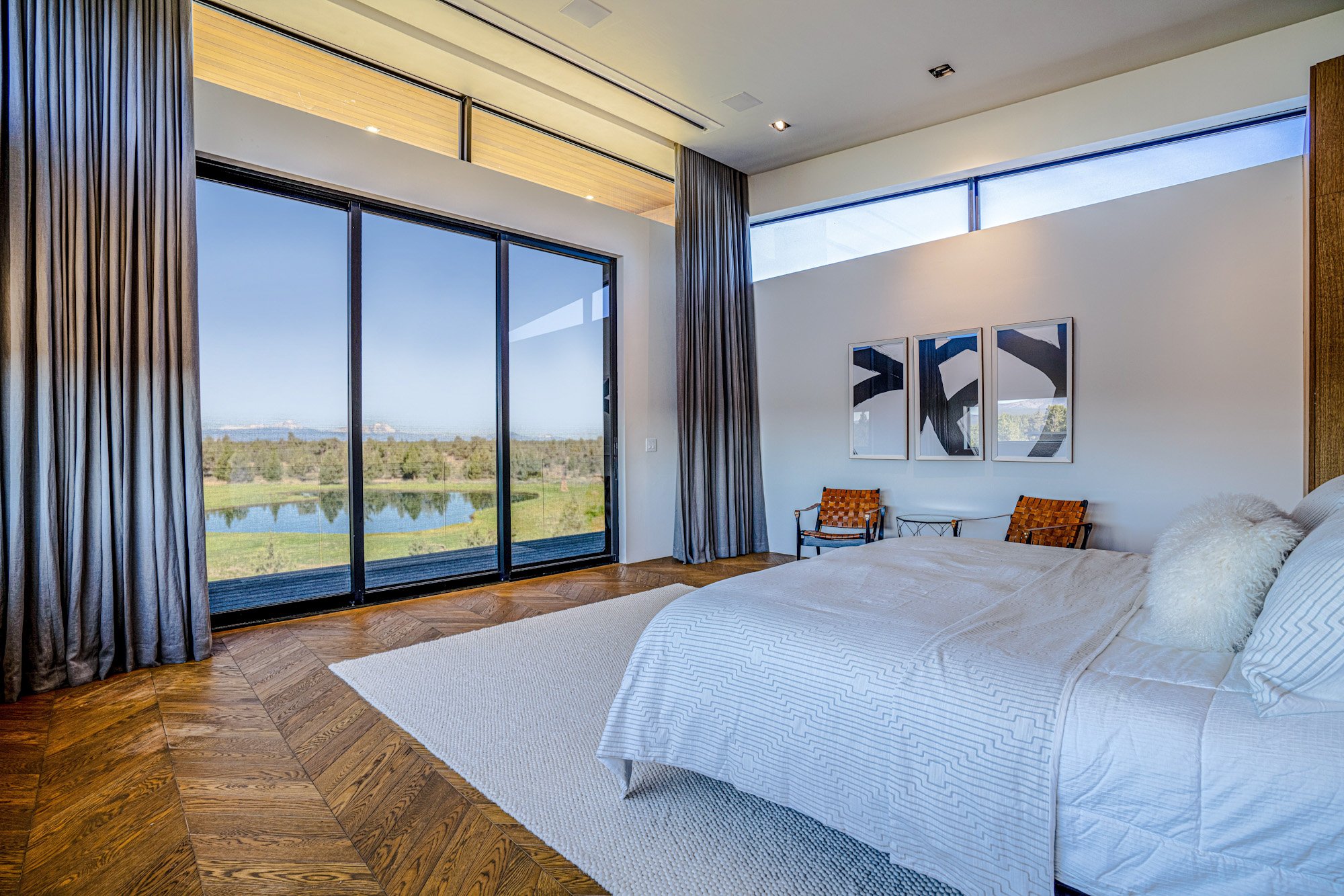 Modern bedroom with a large bed, white bedding, and pillows, next to a big window with a scenic view of a pond, trees, and blue sky.