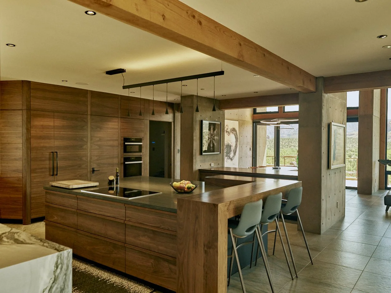 Modern kitchen with wooden cabinets, an island with a dark countertop, and bar stools. Large windows with a view of greenery outside.