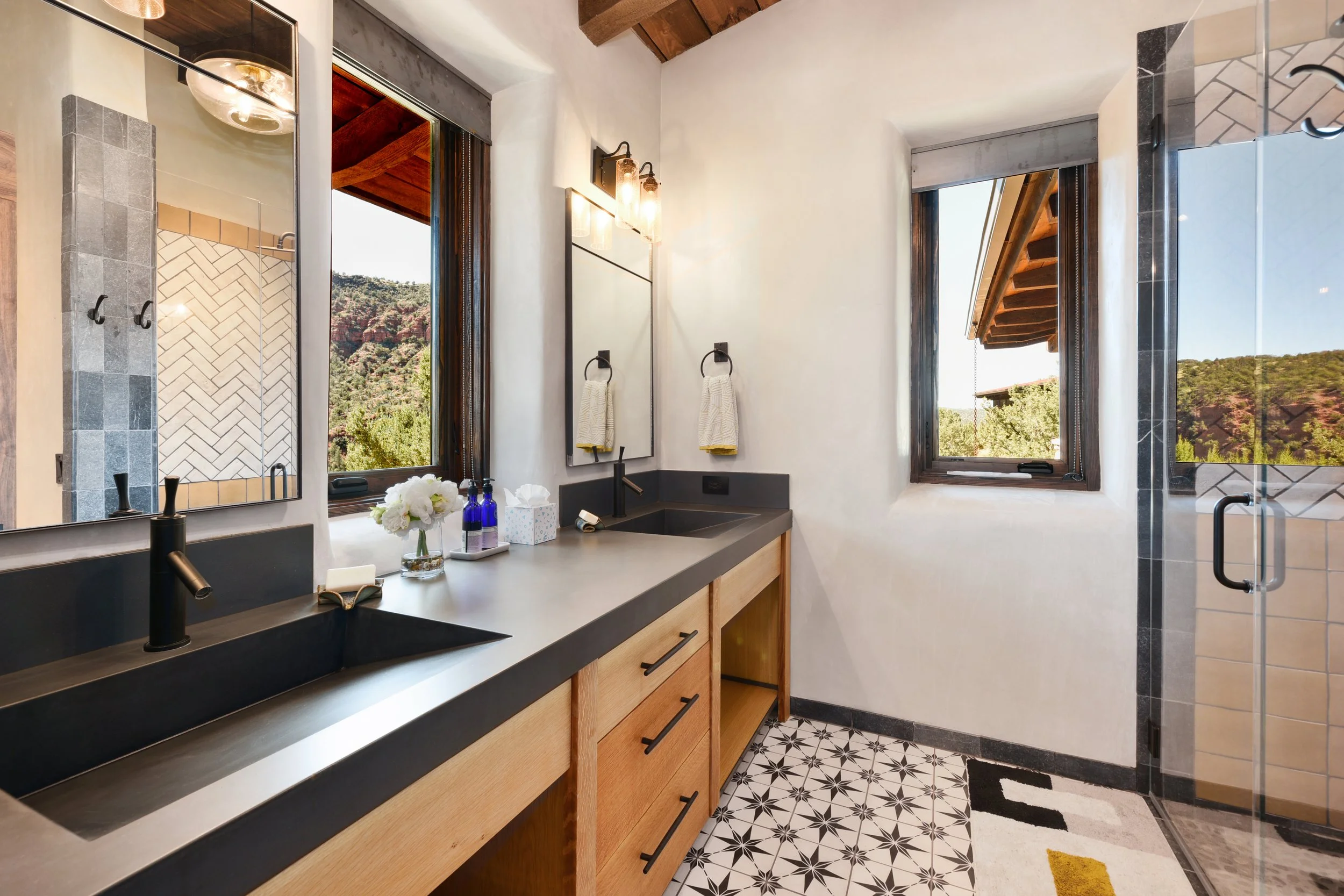 Modern bathroom with dual sinks, black countertops, and a large window with scenic mountain views. Features a patterned tile floor, white walls, and additional small window with a partial view of an outdoor patio.