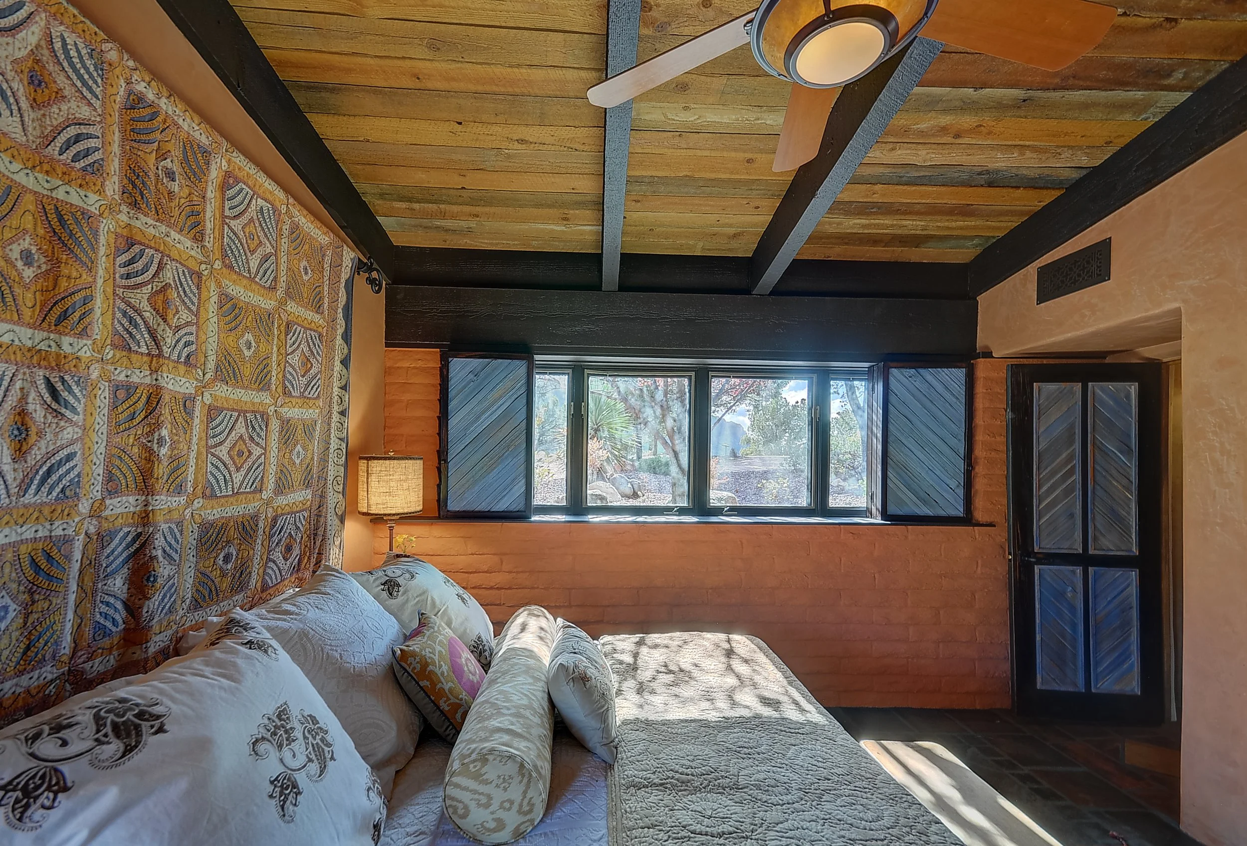 A cozy bedroom with a bed covered in white and beige bedding, decorative pillows, and quilted blanket. There's a nightstand with a lamp beside the bed. A large window with black-framed shutters shows an outdoor scene with trees. The ceiling is rustic wood with exposed beams, and the wall behind the bed has a colorful patterned textile hanging.