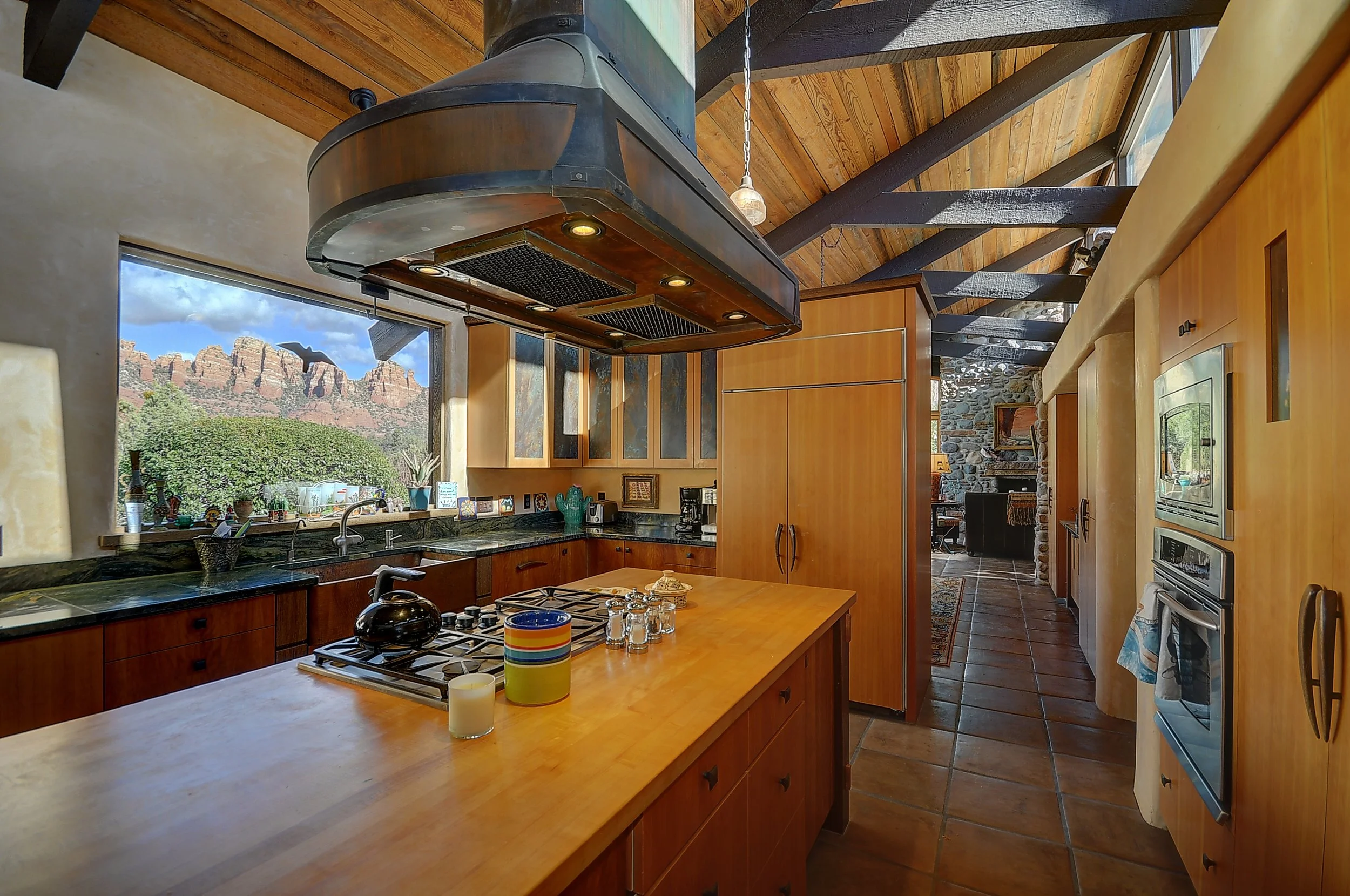 Spacious kitchen with wooden cabinets, black countertops, and a large window showing red rock formations outside. A kitchen island with a kettle and cups, a hood above the stove, and some decorative items on the counter.