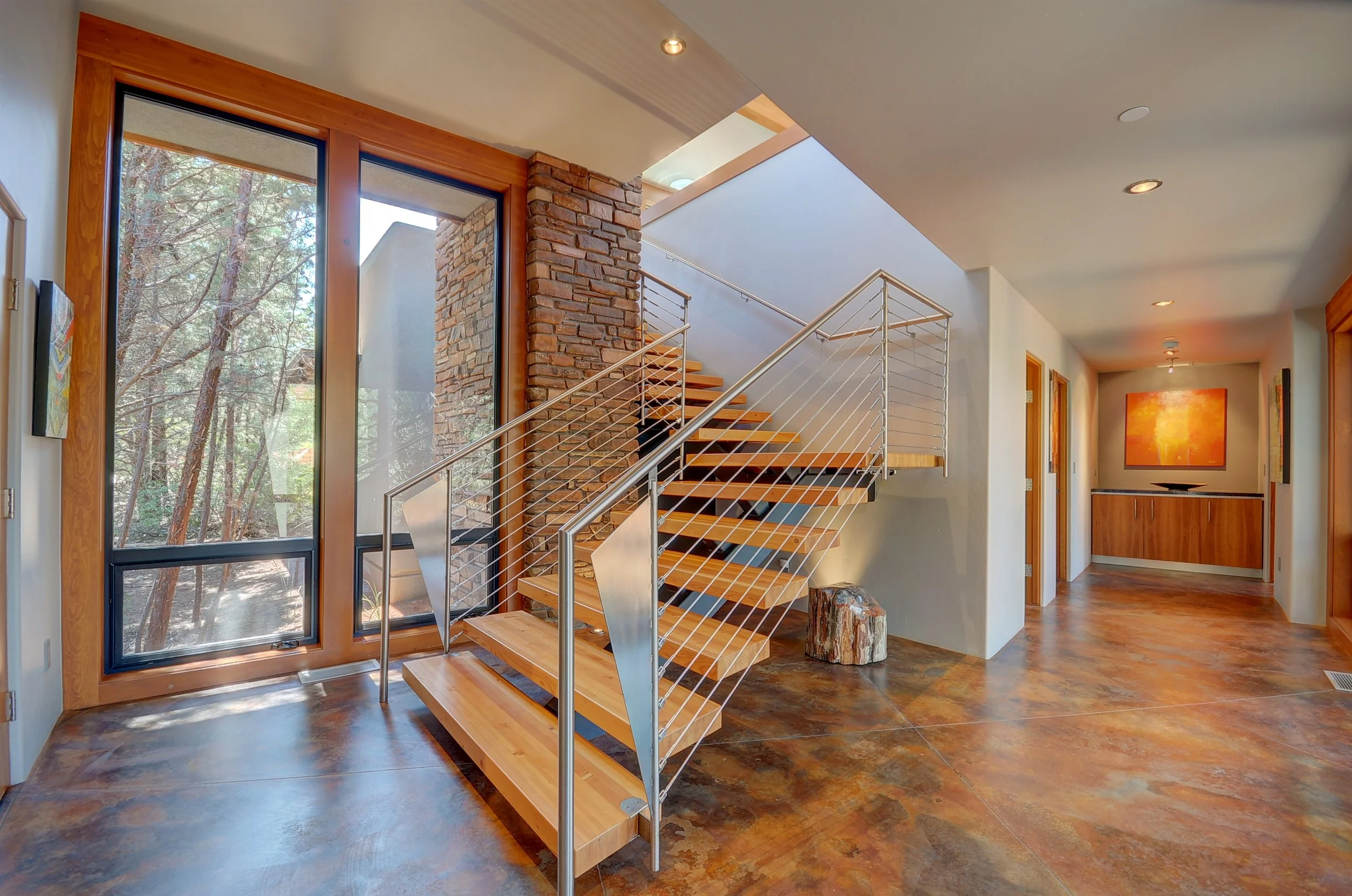 Interior view of a modern home with a staircase, large window with a view of trees, colorful artwork, and warm wooden accents.