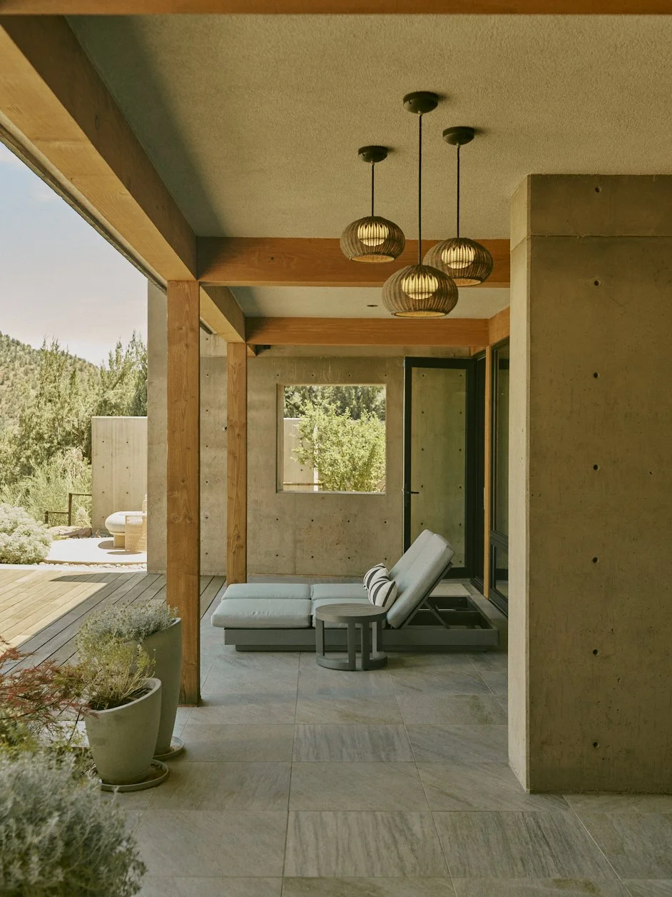 Indoor patio area with lounge chair, small side table, potted plants, wooden beams, modern pendant lights, and sliding glass doors leading outside to a deck with outdoor seating, surrounded by greenery.