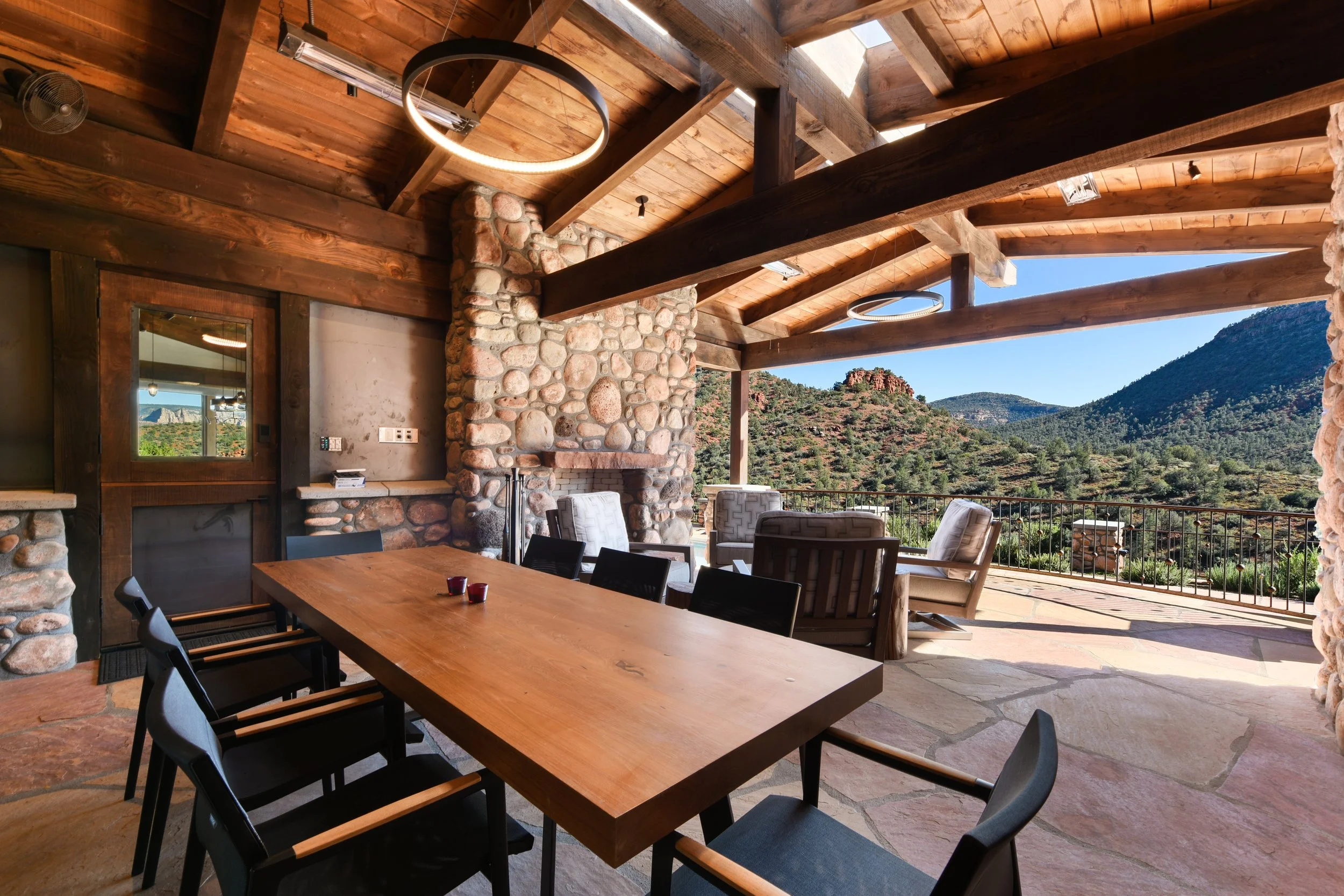 Covered outdoor patio with a large wooden dining table, black chairs, stone fireplace, and seating area with scenic mountain view.