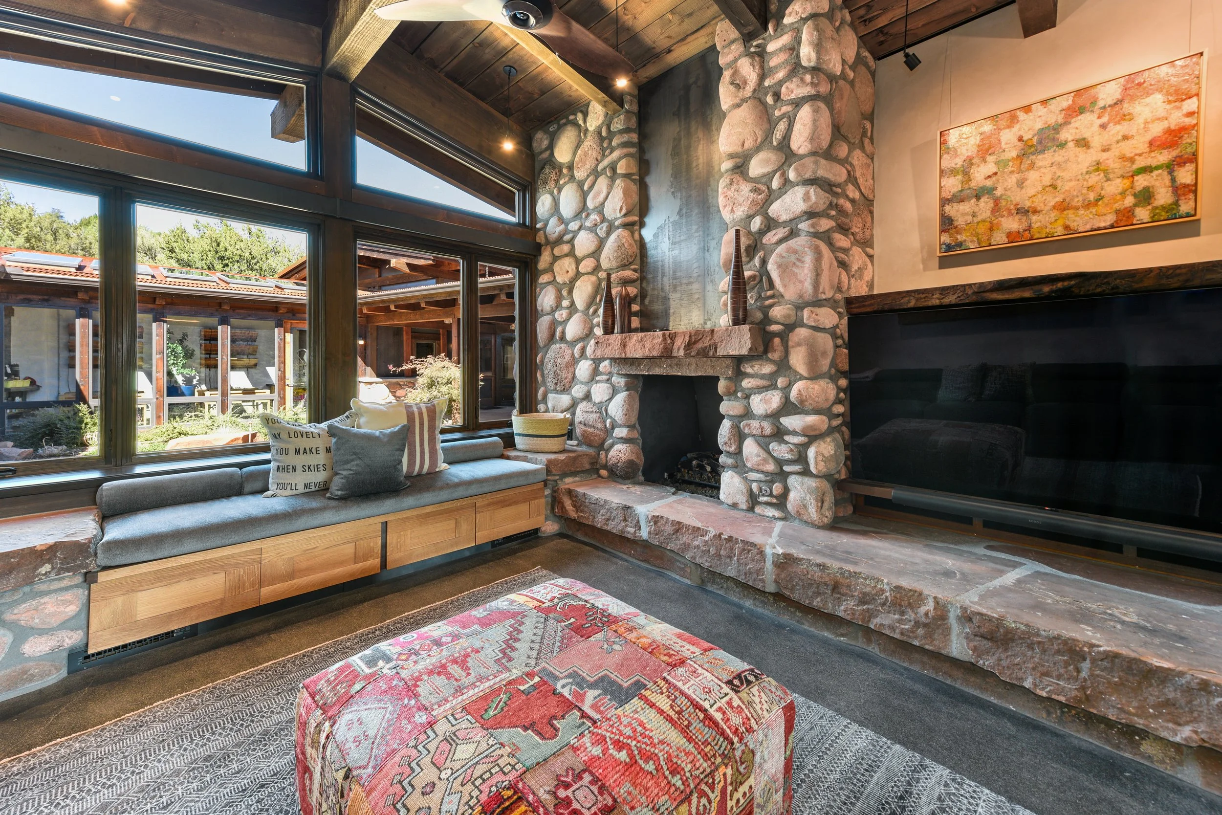 Living room with large windows, stone fireplace, wooden ceiling, colorful abstract painting, cushioned window seat with pillows, patterned ottoman, and sofa.