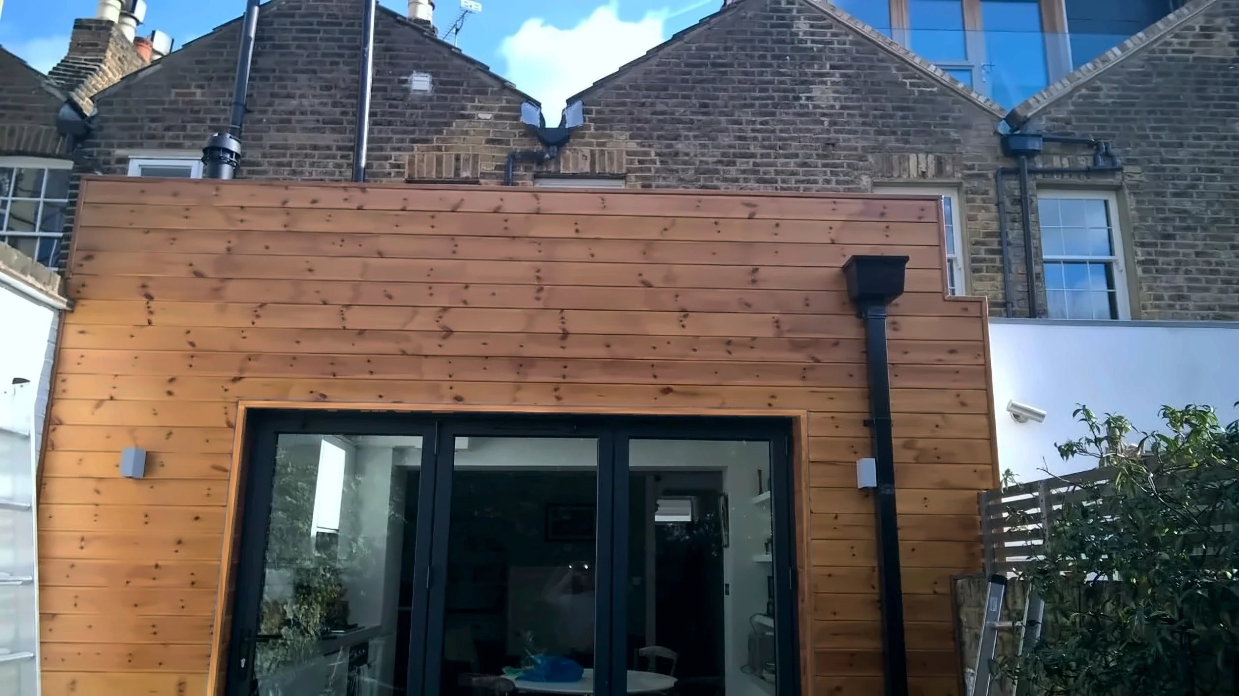 Full-width rear extension with natural timber cladding and black aluminium bifold doors at a rear kitchen extension on Short Road, Chiswick, W4 by Robin Services.