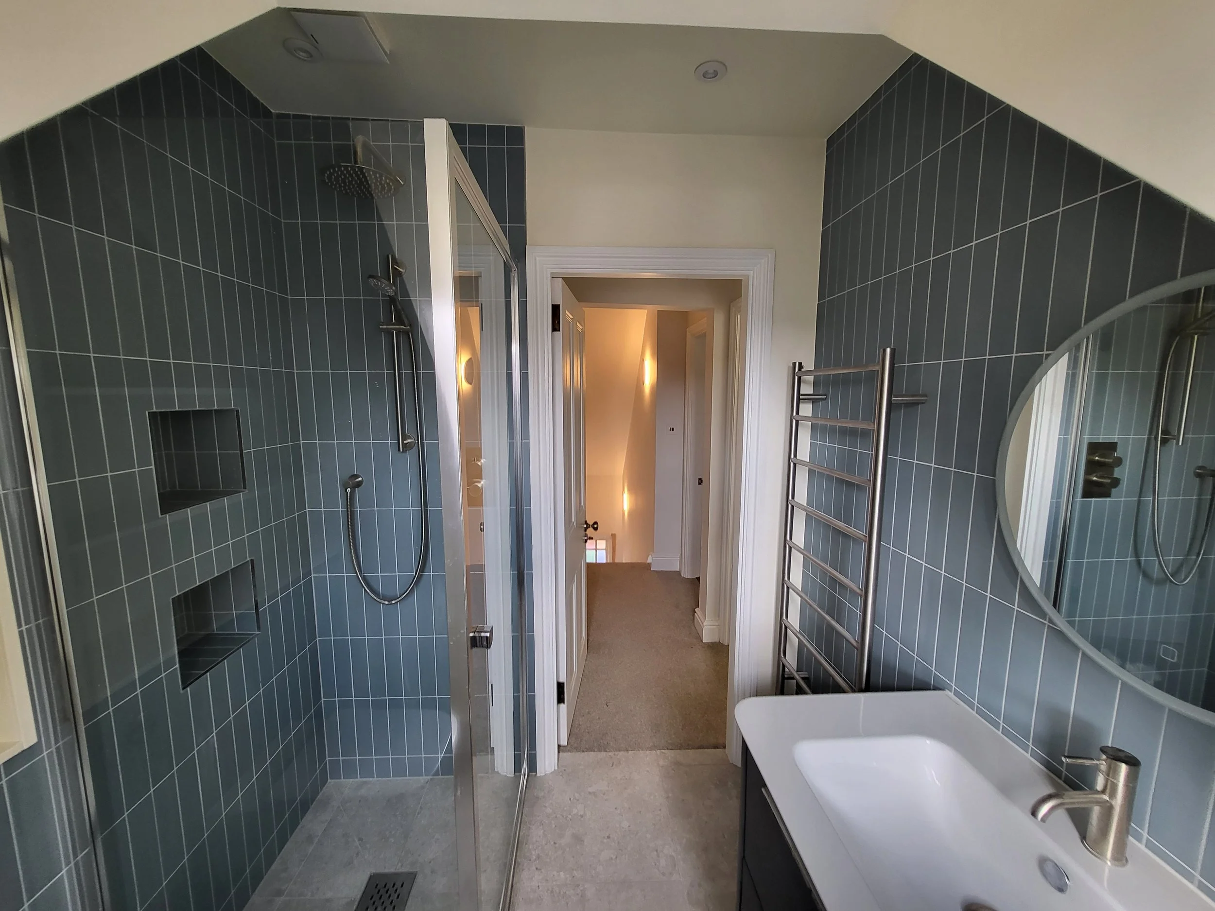 Modern bathroom with a walk-in shower featuring dark green tiles and built-in shelves, a white sink with a gold faucet, a large round mirror, a towel radiator, and a view into a hallway with carpeted floor.