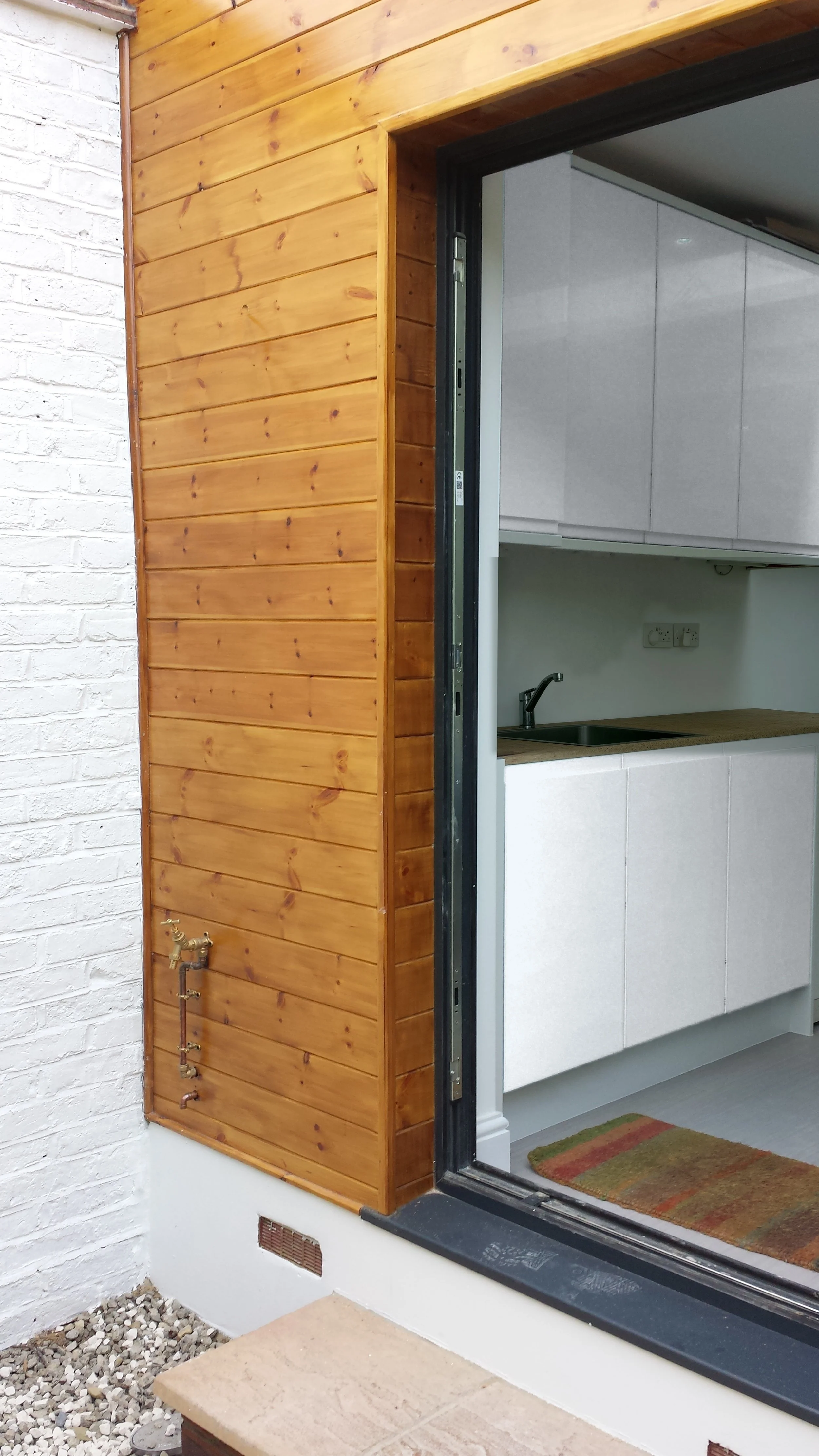 Open side door in natural pine cladding showing white gloss kitchen units and worktop, at a rear kitchen extension on Short Road, Chiswick, W4 by Robin Services.